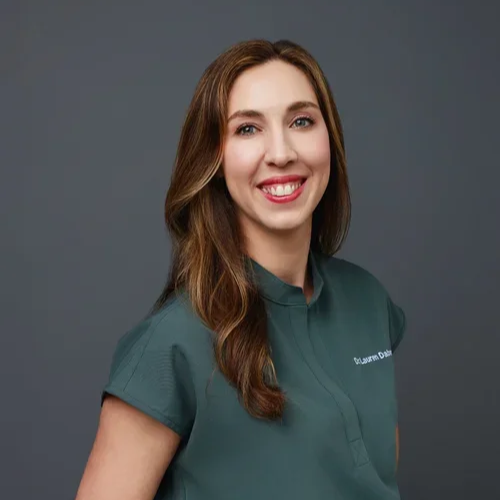 A young woman with long brown hair, wearing a teal medical uniform, smiling against a plain gray background.