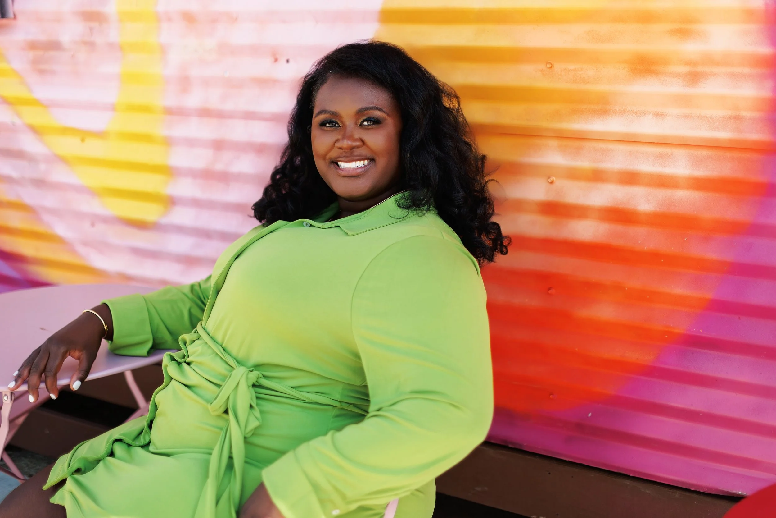 A woman with dark curly hair, smiling, wearing a bright green dress, sitting on a pink chair in front of a colorful, striped background.