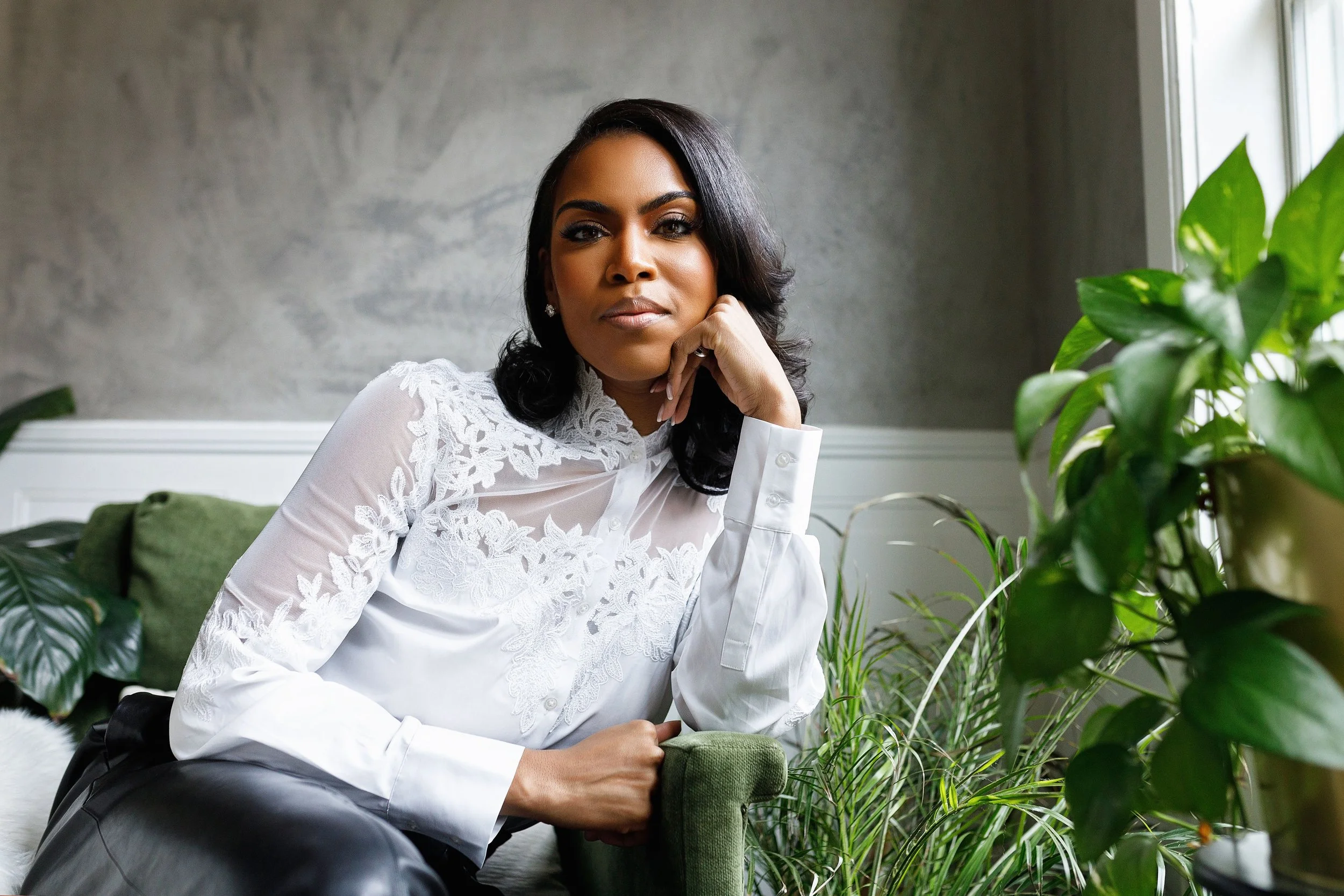 A woman with dark hair and a white embroidered blouse sitting on a green armchair near plants in a room with gray walls and a large window.