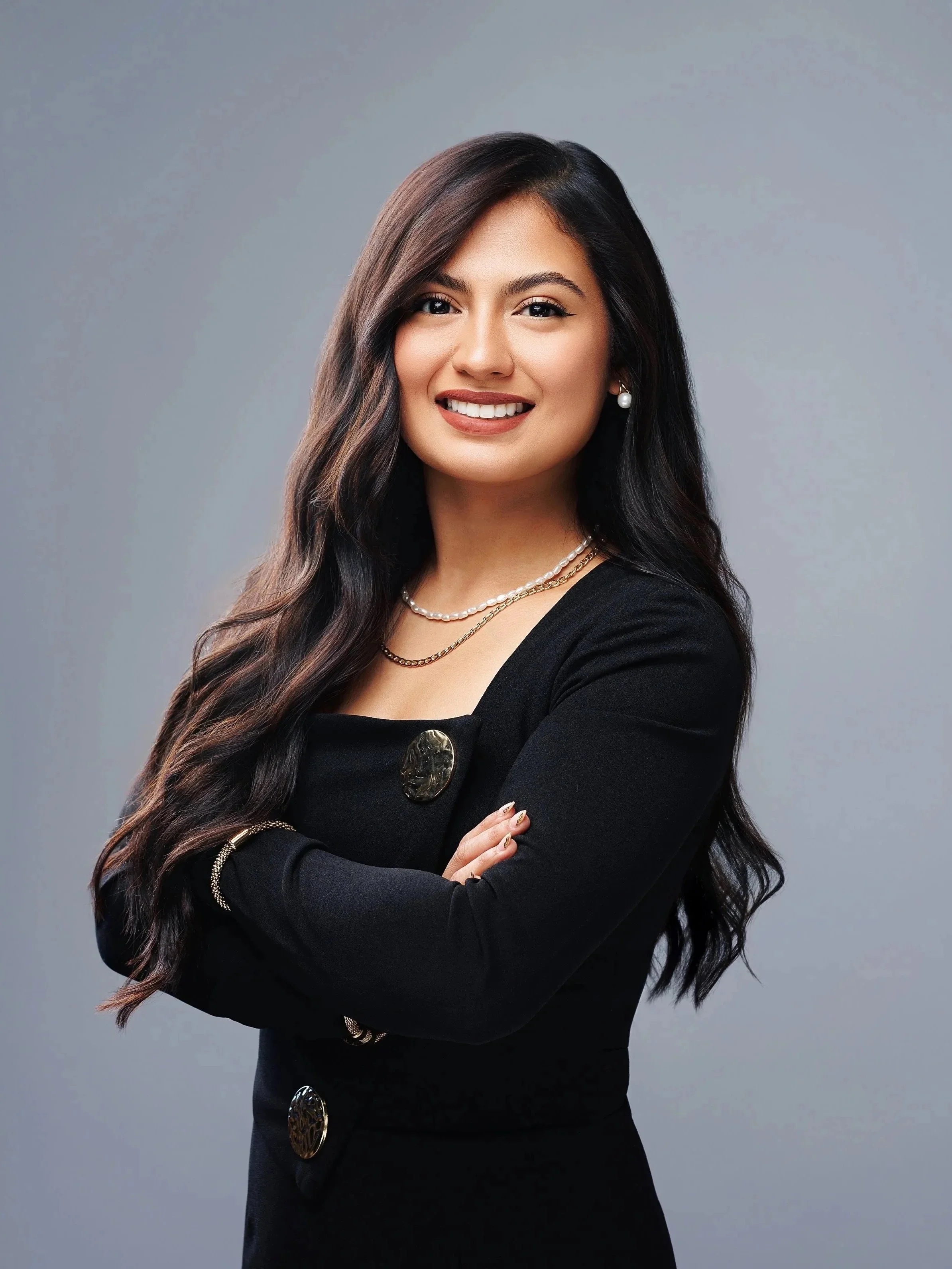 A woman with long dark hair smiling, crossing her arms, dressed in black with decorative buttons, wearing pearl earrings, layered necklaces, and a bracelet, posing against a gray background.