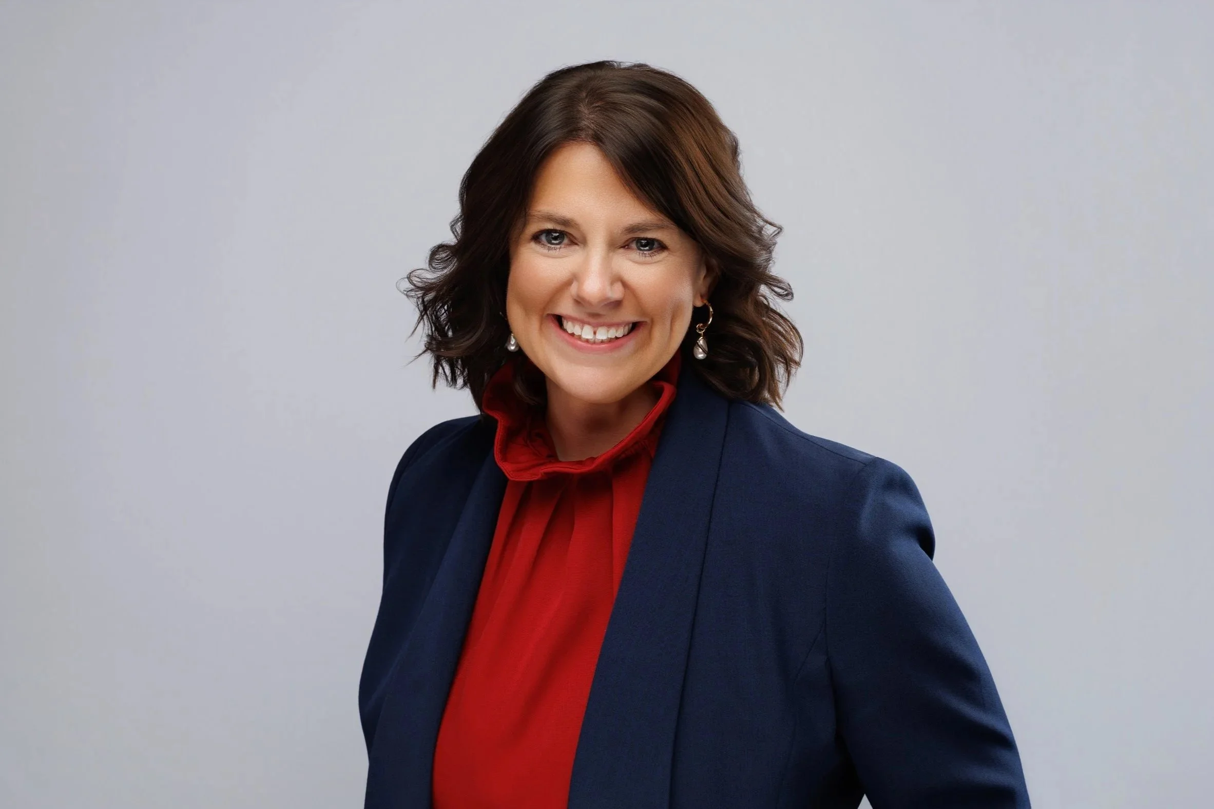 A woman with shoulder-length dark brown hair and a bright smile, wearing a navy blazer and a red blouse with a ruffled neckline, posing against a plain light gray background.