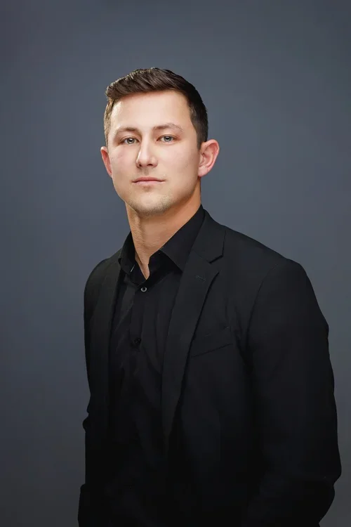 A young man with short brown hair and a serious expression, wearing a black suit and black shirt, standing against a gray background.