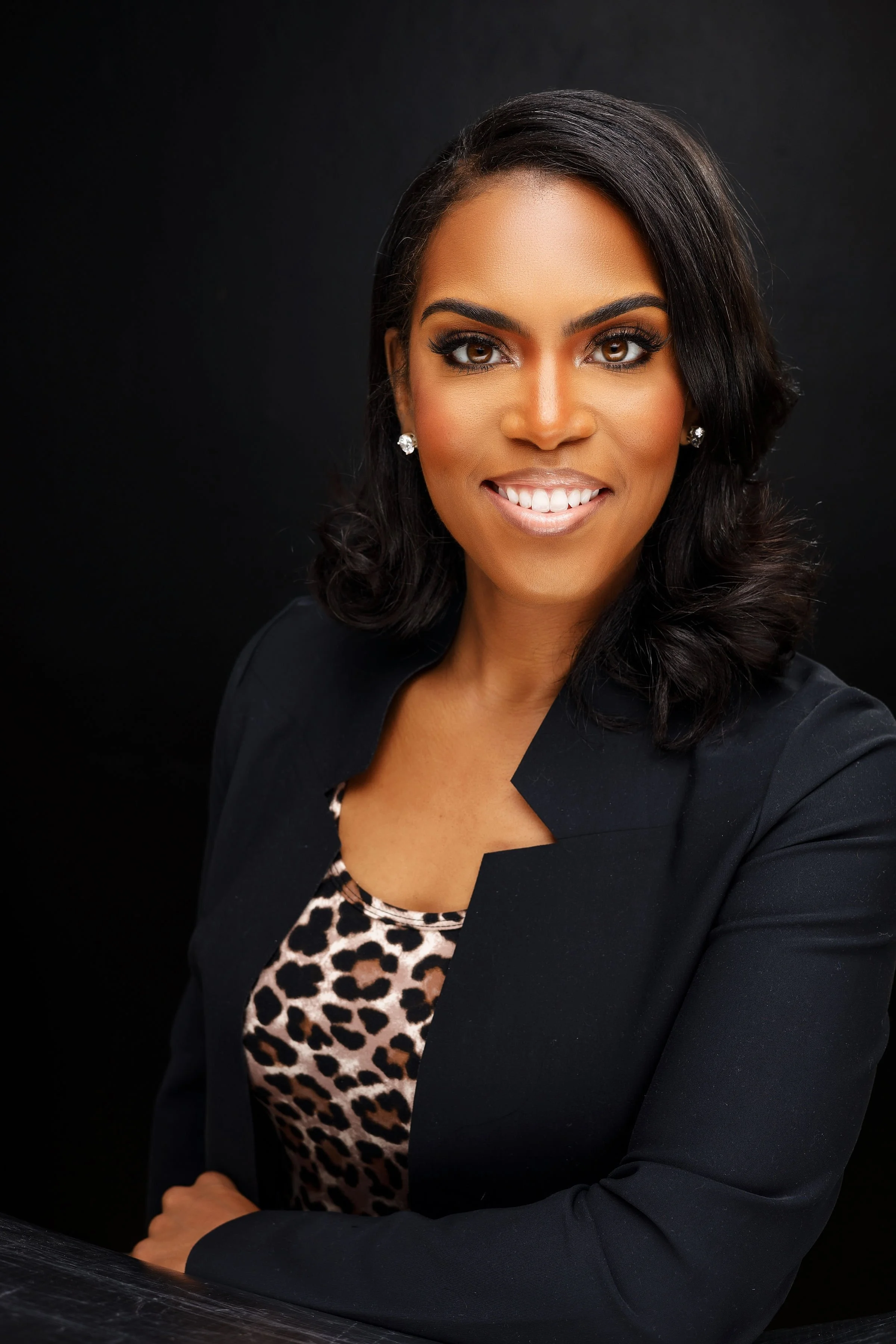Professional woman with dark hair, wearing a black blazer and leopard print top, smiling against a black background.