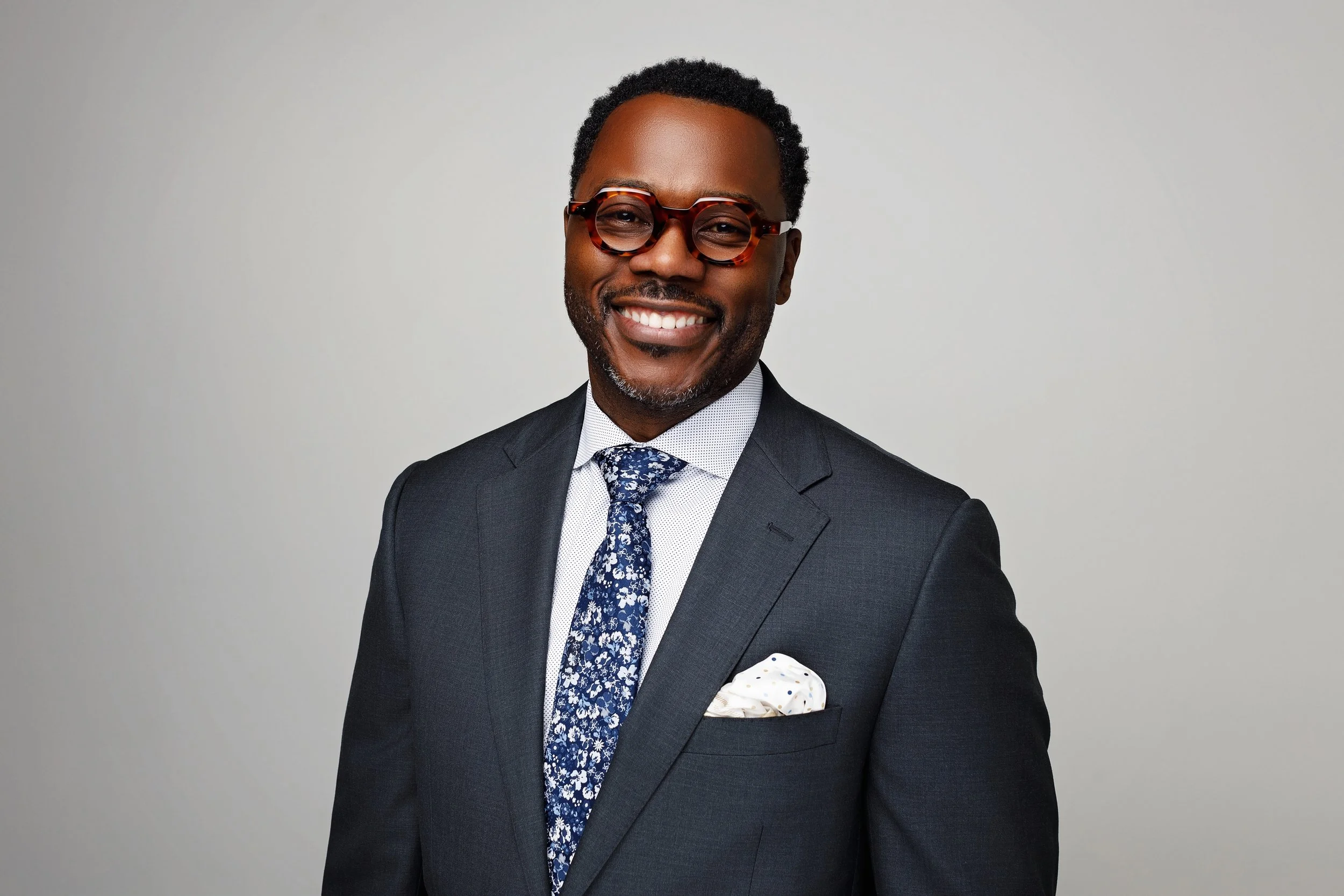 A smiling African American man in a dark suit, white shirt, patterned blue tie, and tortoise-shell glasses, standing against a light gray background.