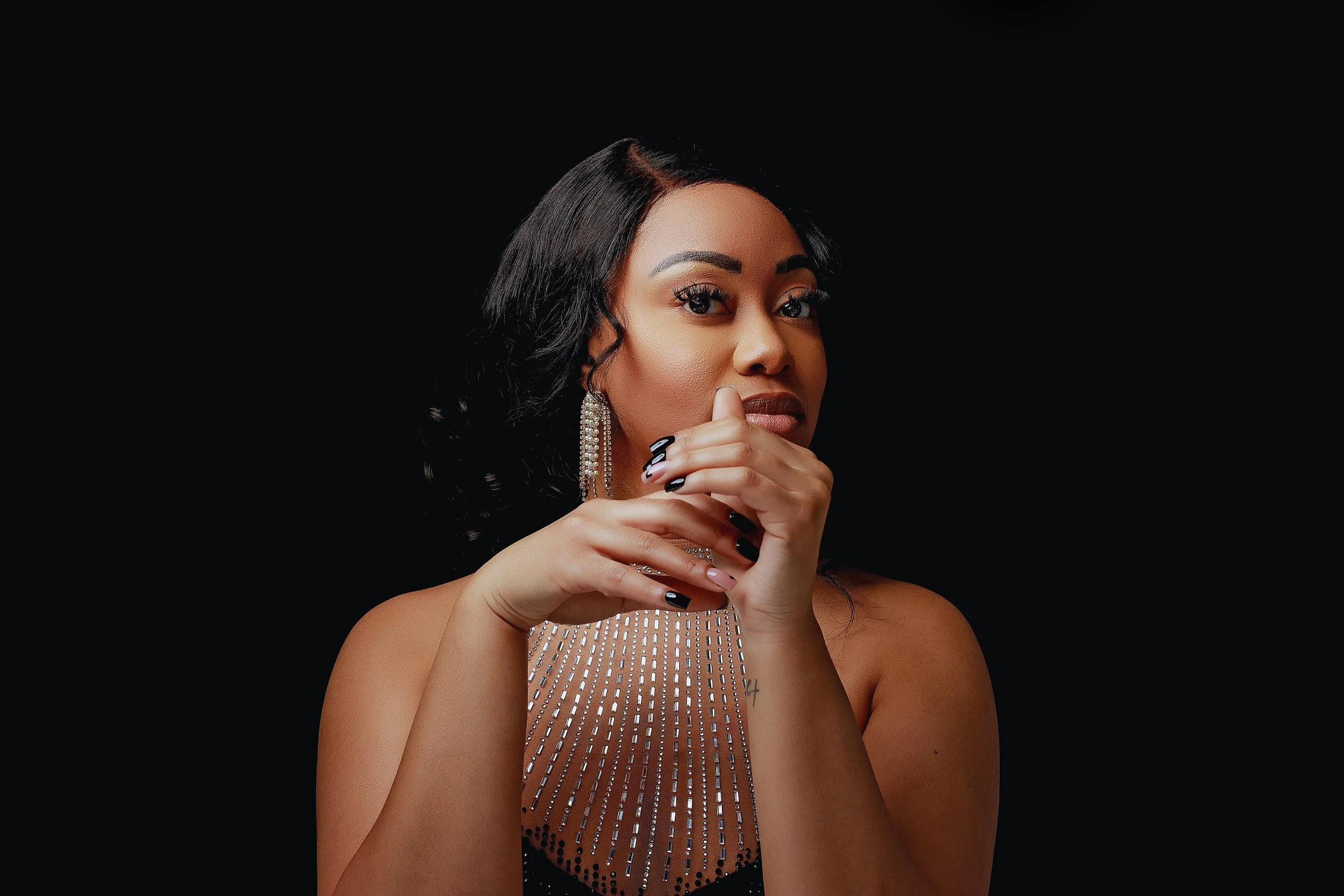 Portrait of a woman with dark hair and makeup, wearing a beaded dress and long earrings, against a black background.