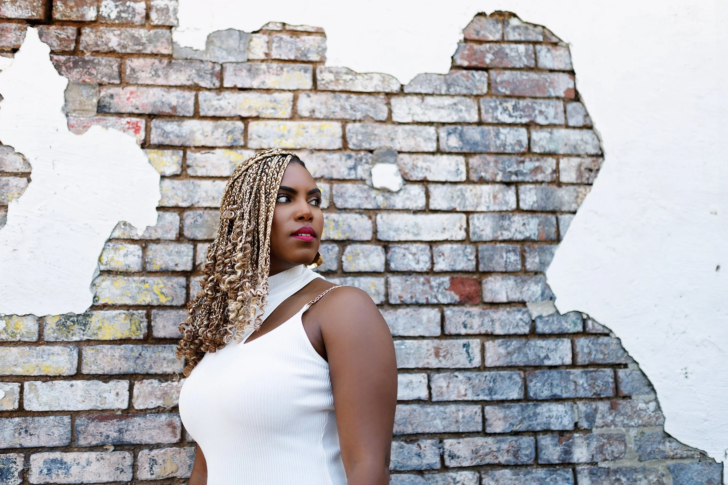 A woman with blonde and black braided hair stands against a worn brick wall with a partially painted white wall, looking to the side.