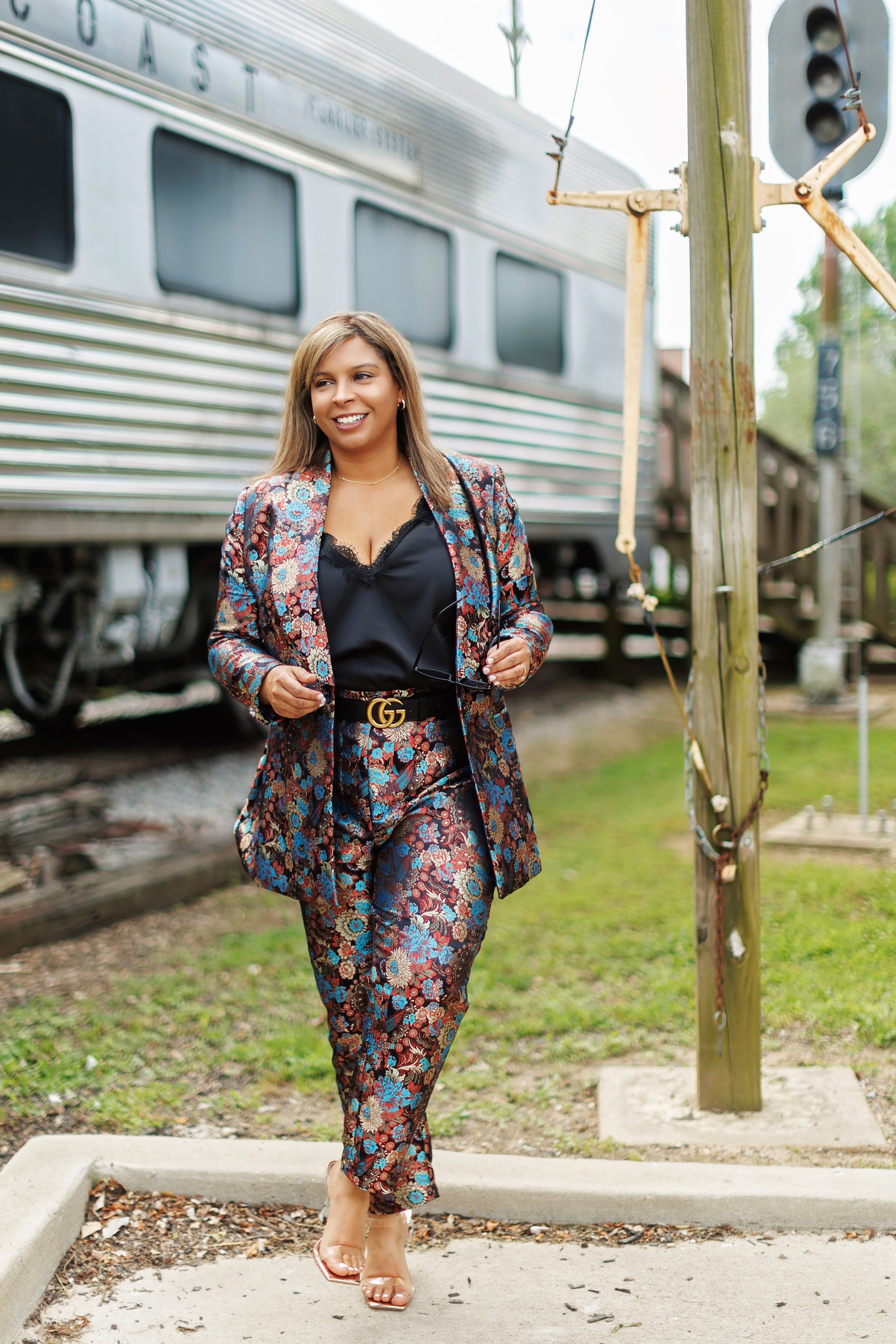 A woman with long hair walking outdoors near a train, wearing a floral patterned pantsuit, black top, clear heels, and a Gucci belt, holding sunglasses, smiling.