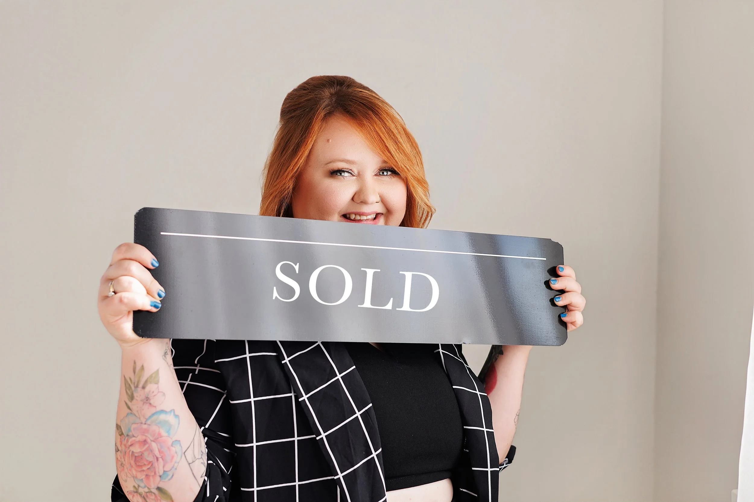 Woman with red hair, tattoos, and a checkered blazer holding a sign that says 'SOLD'.