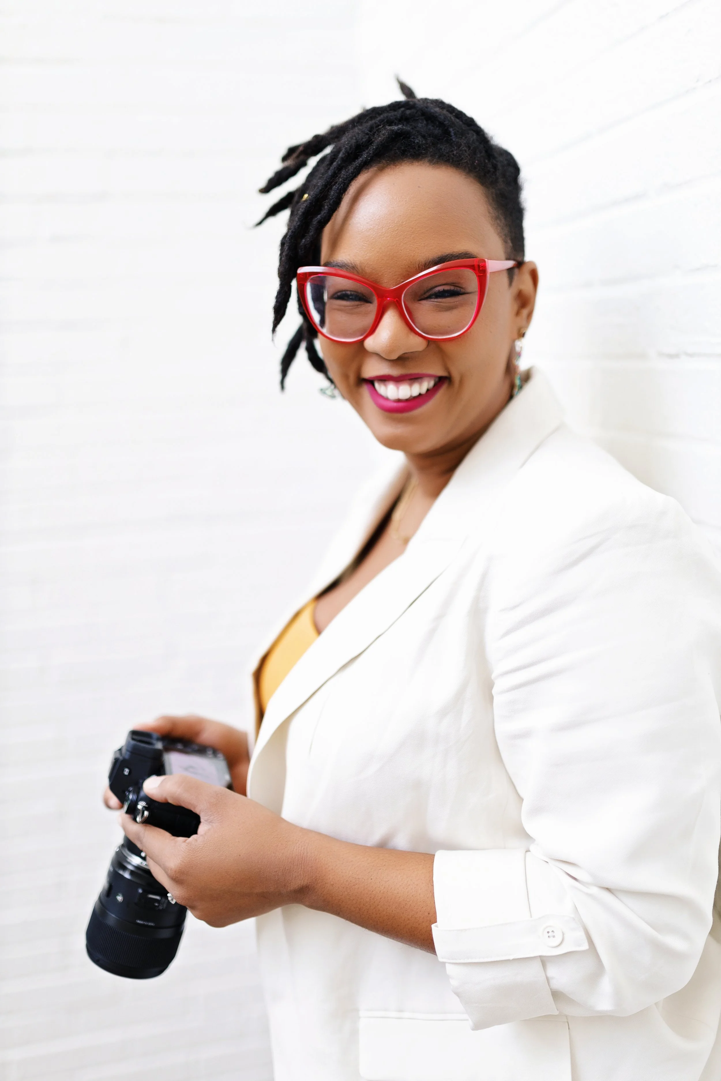 A woman with dreadlocks wearing red glasses and a white blazer, smiling, holding a camera, standing against a white brick wall.