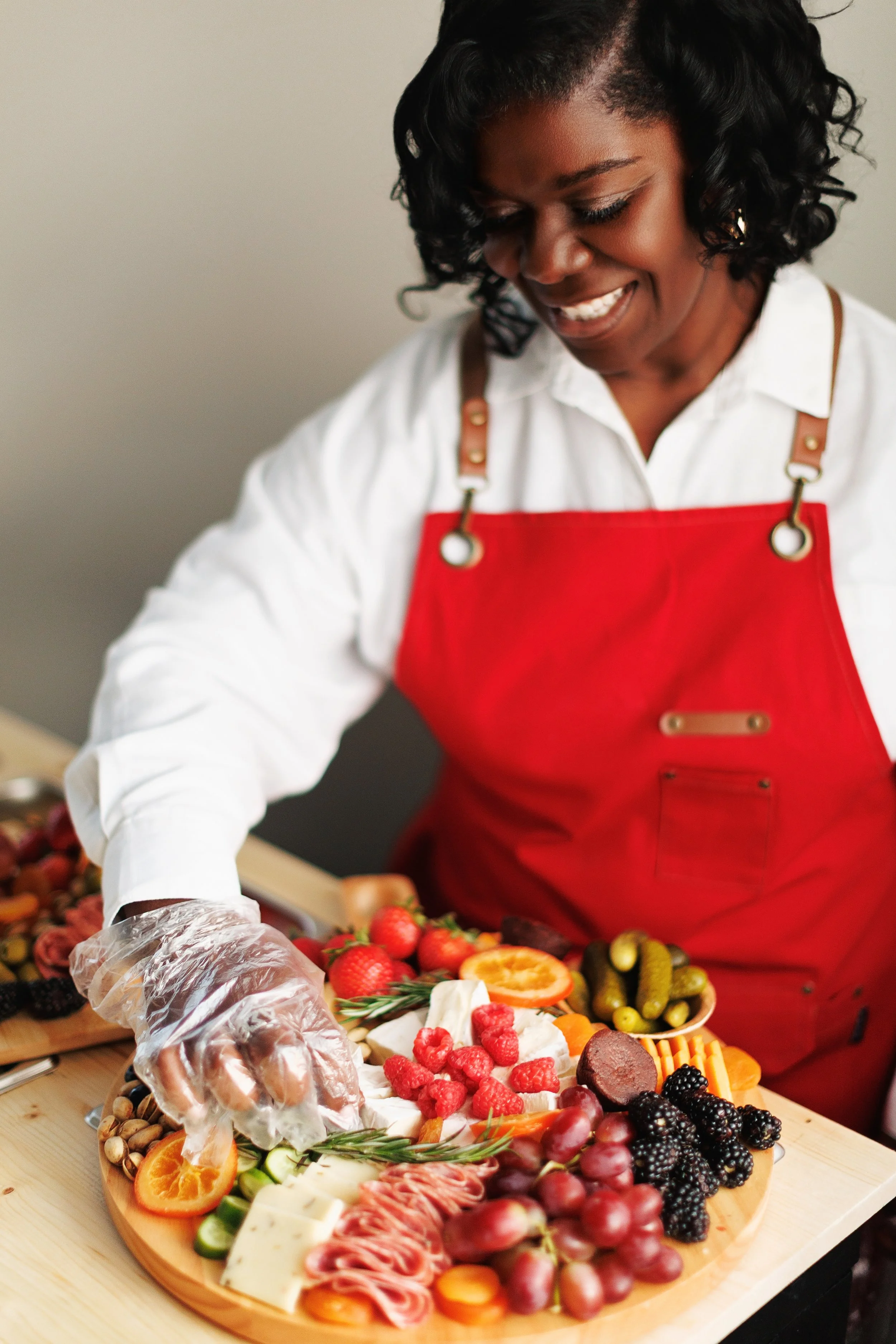 A woman wearing a red apron and white shirt arranging a cheese and fruit platter with strawberries, blackberries, grapes, orange slices, cucumbers, and meats.