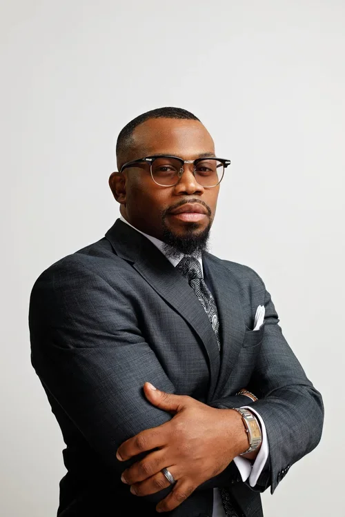 A professional African American man in a dark suit, with glasses, arms crossed, standing against a plain white background.