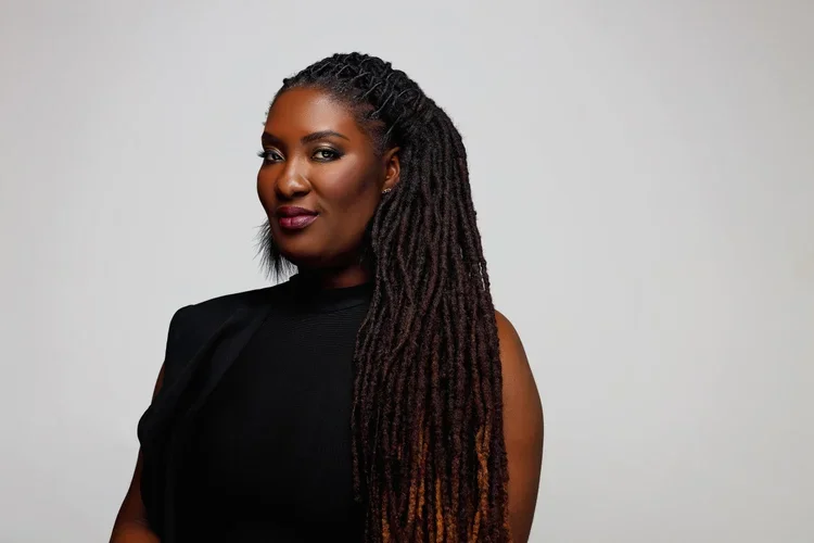 A woman with dark skin and long dreadlocks wearing a black sleeveless top, posing confidently against a plain gray background.