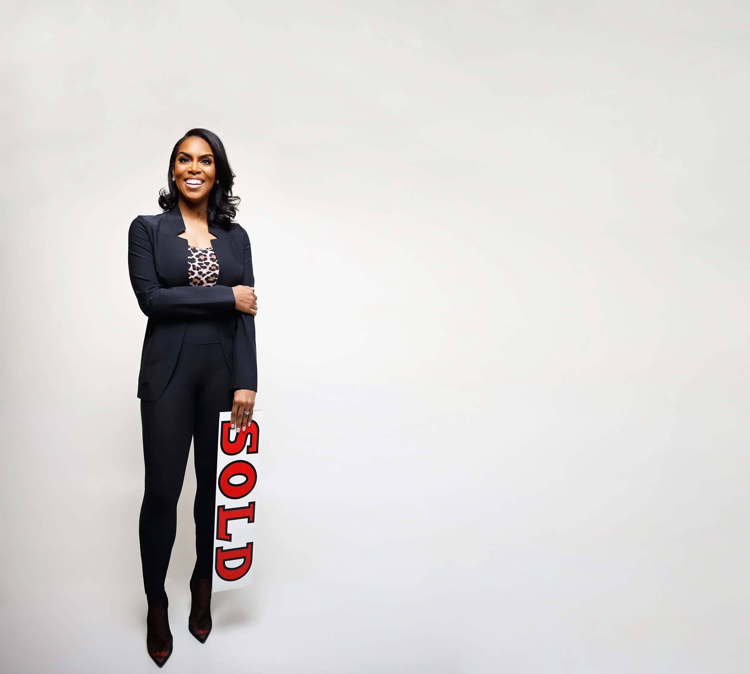 A woman dressed in a black suit with a leopard print top underneath, standing against a plain light-colored background, holding a sign that reads 'STOP' in red and white.