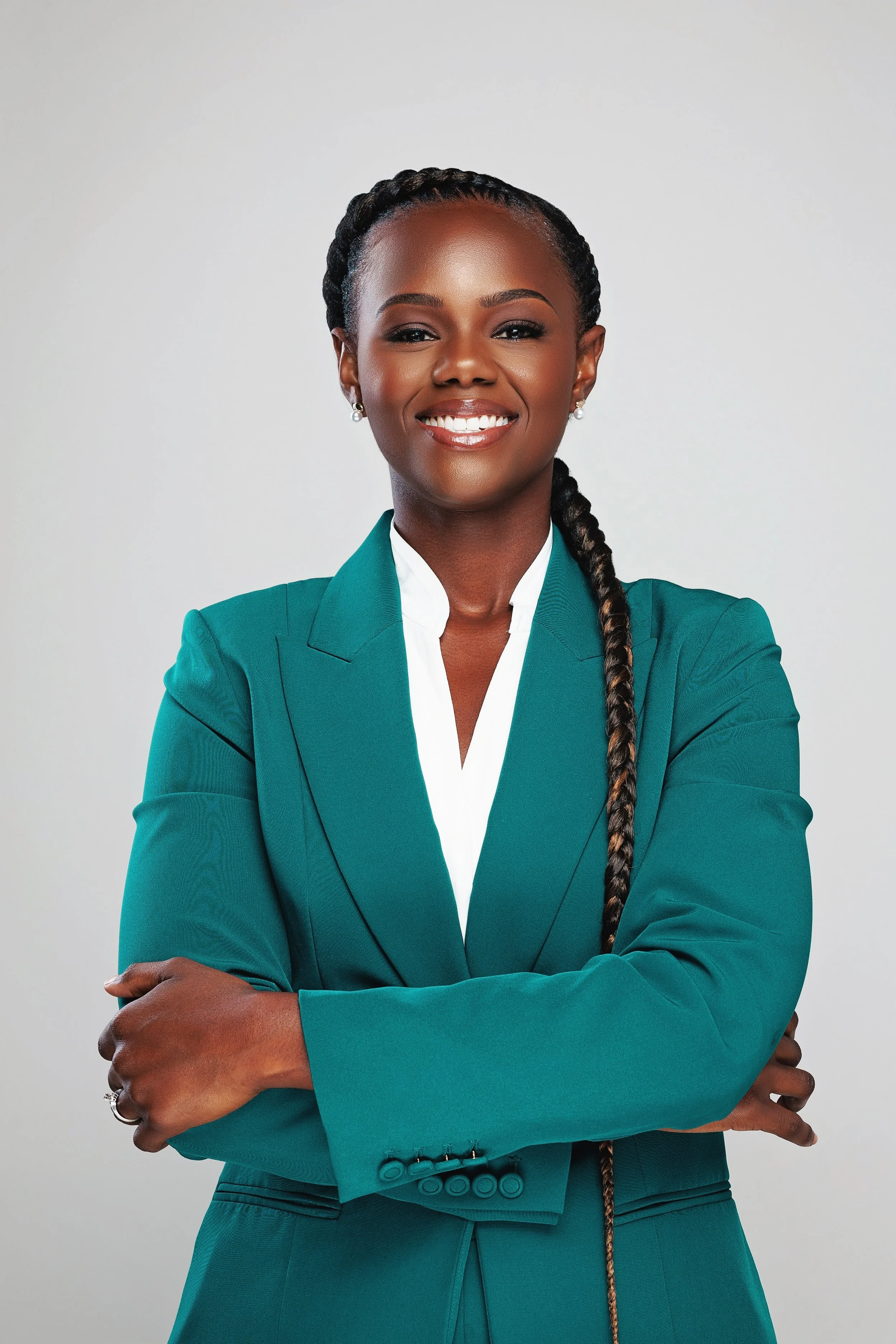 A confident woman with dark skin, wearing a teal business suit and white shirt, standing with arms crossed and smiling.