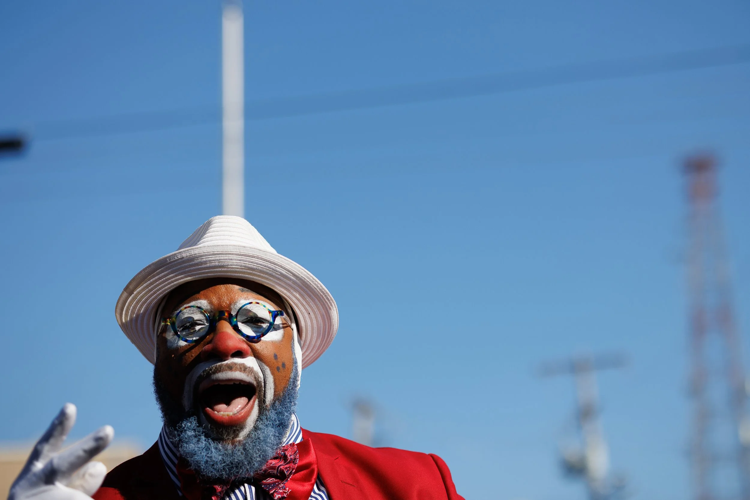A man wearing colorful face paint, round glasses, a white hat, a red blazer, and a striped blue and white shirt, joyfully smiling and making a peace sign with his hand, outdoors under a clear blue sky.