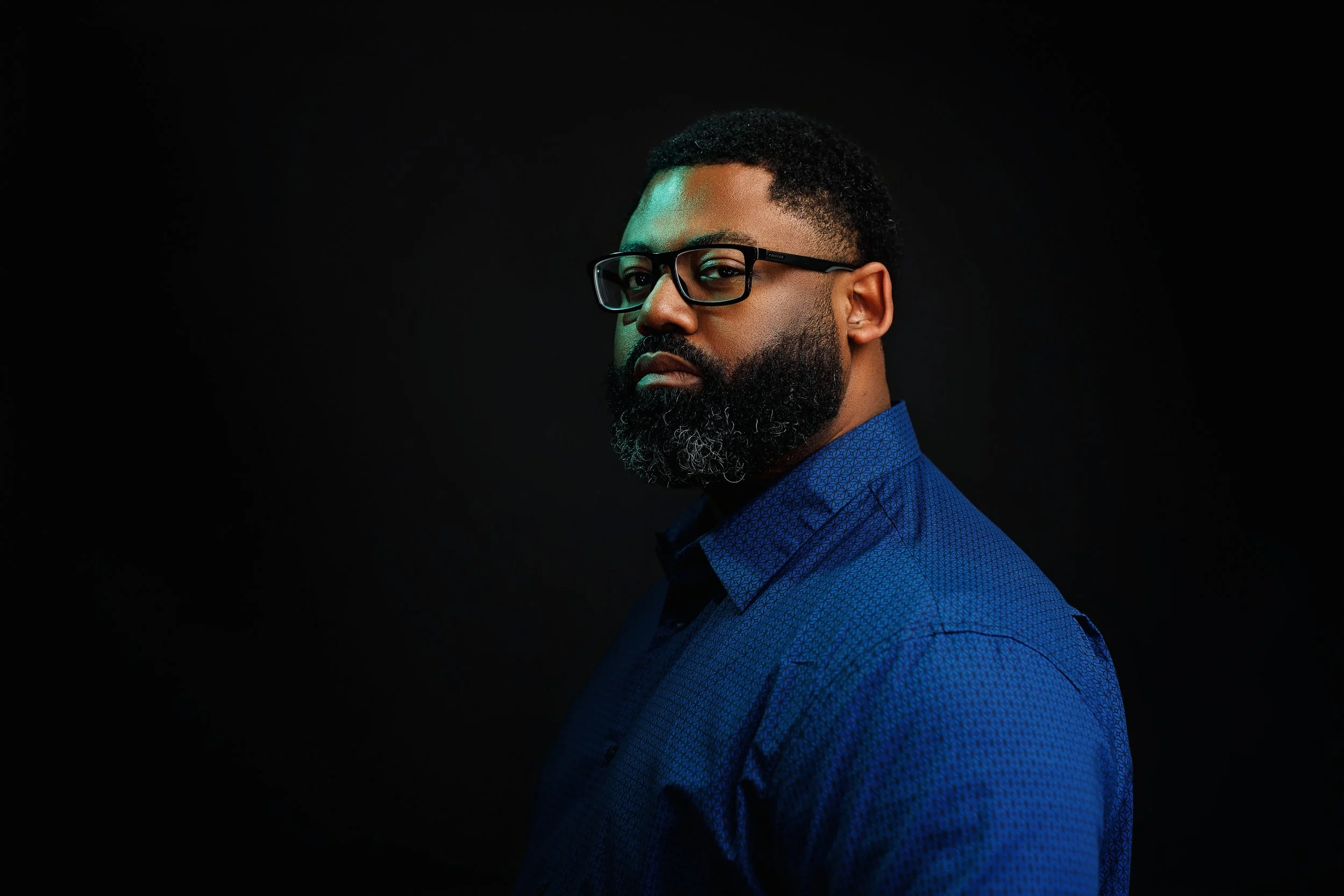 Portrait of a man with glasses, a beard, and short curly hair, wearing a blue shirt against a dark background.