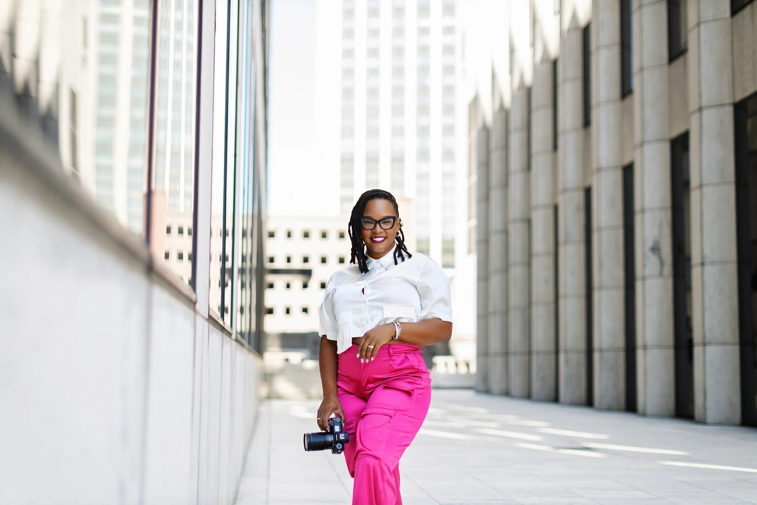 A woman with glasses, dark lipstick, and dreadlocks walking on a city sidewalk holding a camera, with tall modern buildings in the background.
