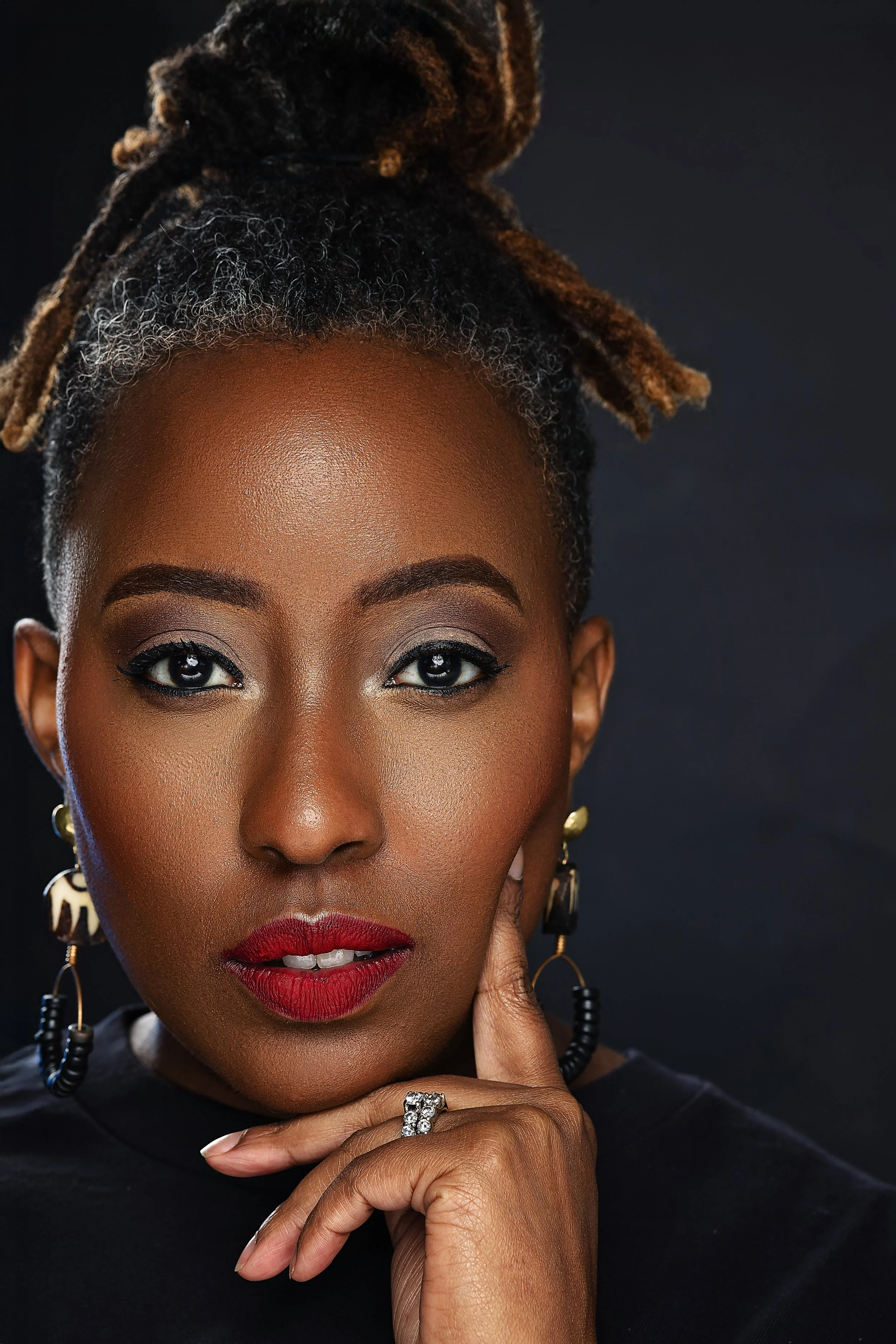 Close-up portrait of a Black woman with styled dreadlocks, wearing bold red lipstick, eye makeup, and large earrings, touching her chin with her hand and looking at the camera against a dark background.