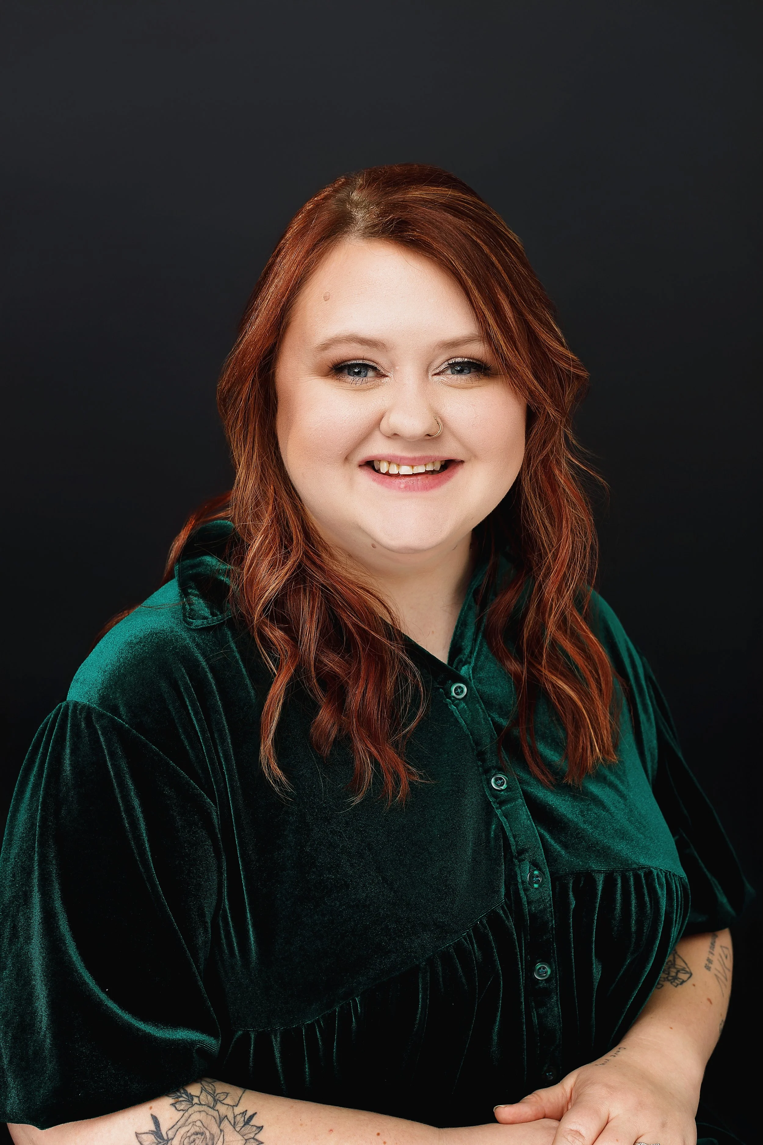 A woman with red hair and a nose ring smiling at the camera, wearing a dark green velvet shirt with tattoos visible on her arm against a black background.