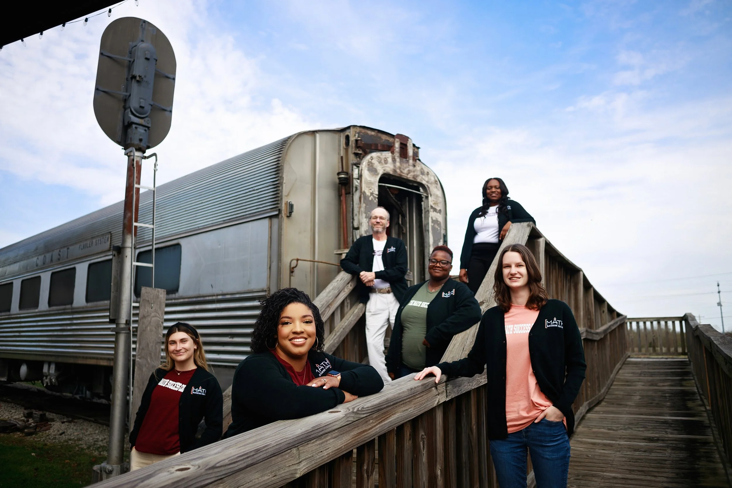 Group of six diverse people standing on a wooden outdoor staircase next to a vintage train car, some smiling and looking at the camera. The sky is partly cloudy.