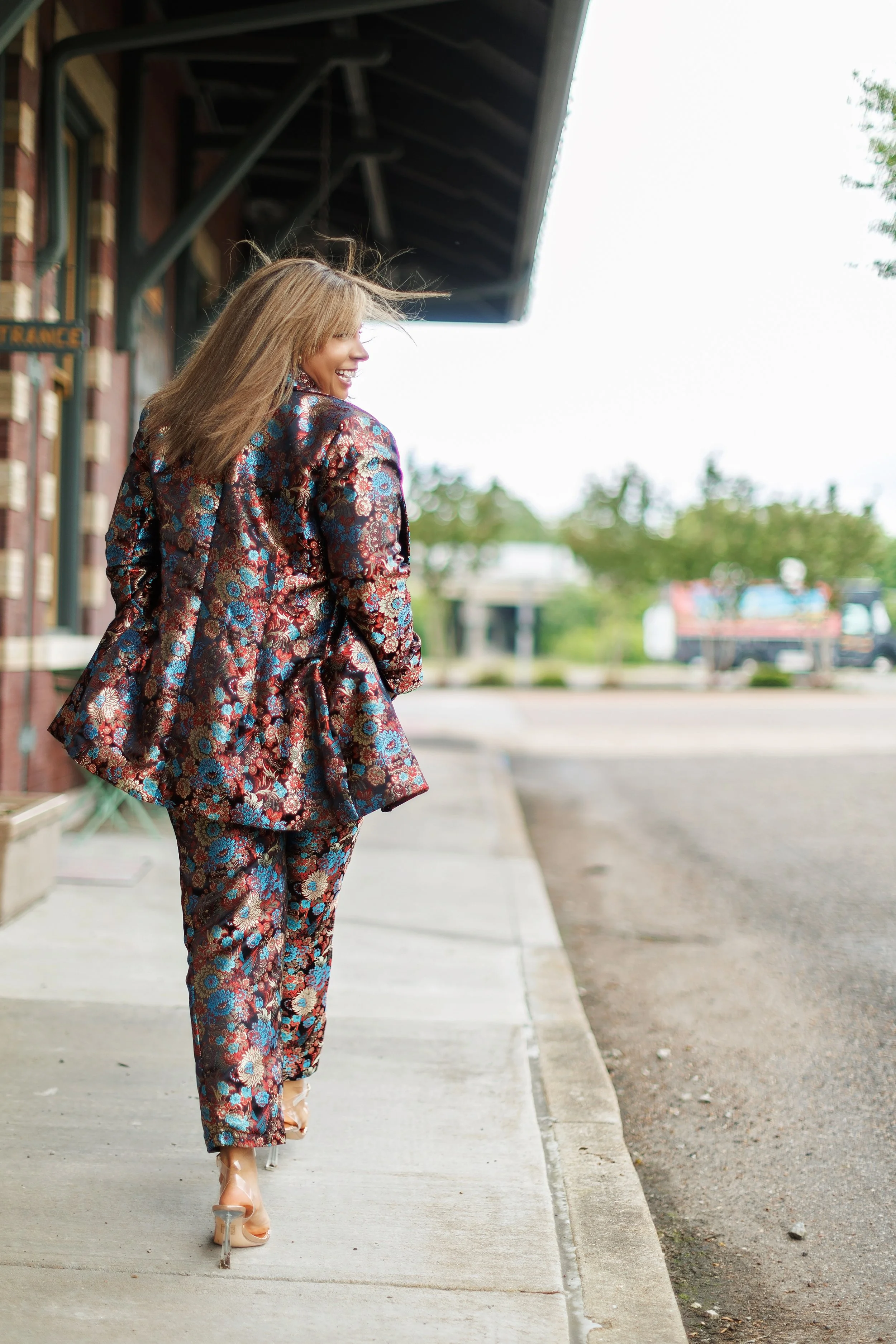 Woman in a floral suit smiling and walking on the sidewalk outside a brick building.