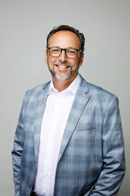 A smiling middle-aged man with glasses, wearing a light blue plaid blazer and white shirt, standing against a plain gray background.