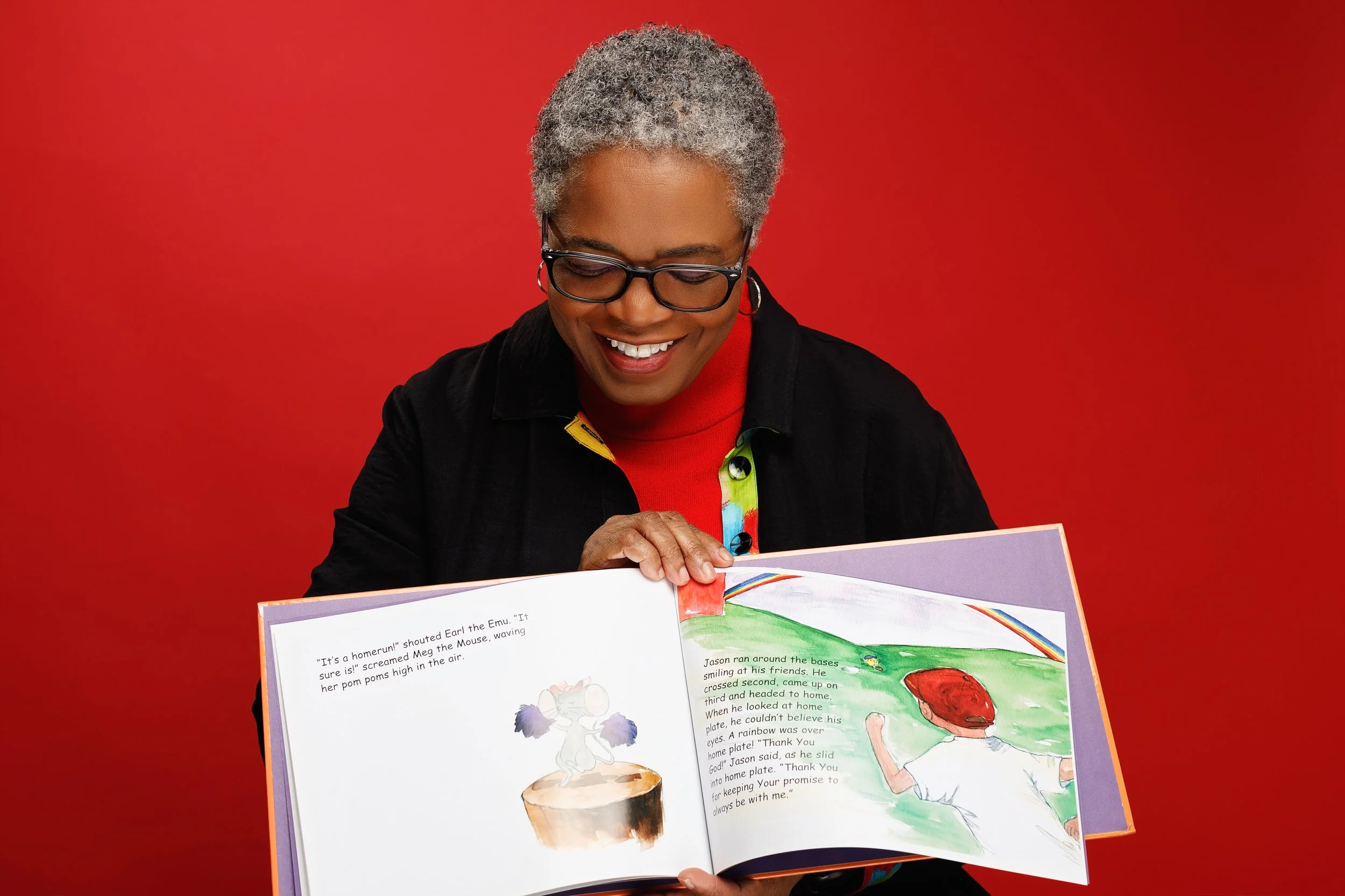 An elderly woman with gray curly hair, glasses, and earrings, smiling while holding and reading a children's picture book against a red background.