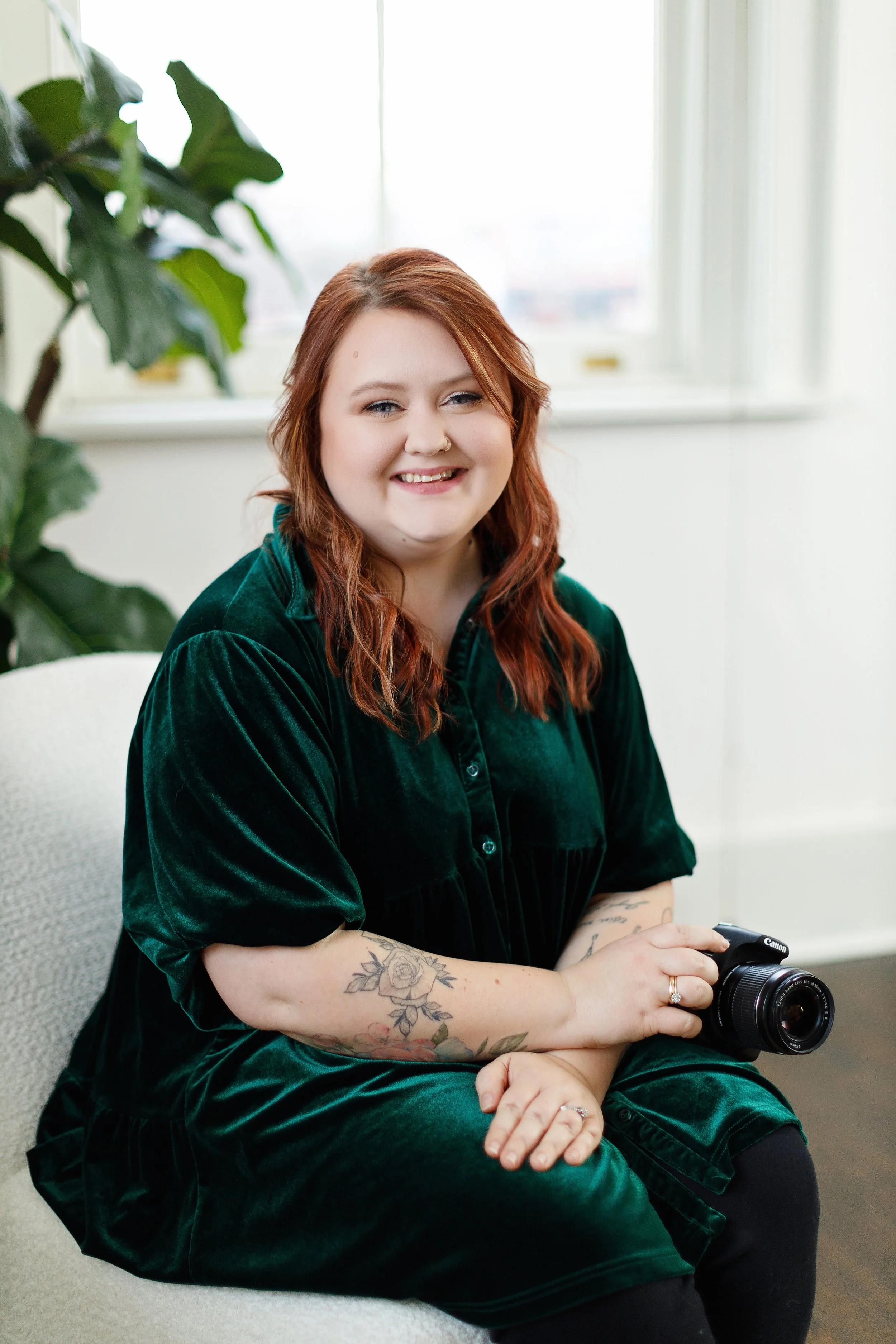 A woman with red hair and tattoos, sitting on a beige armchair, holding a camera, smiling indoors with a large window and green plant in the background.