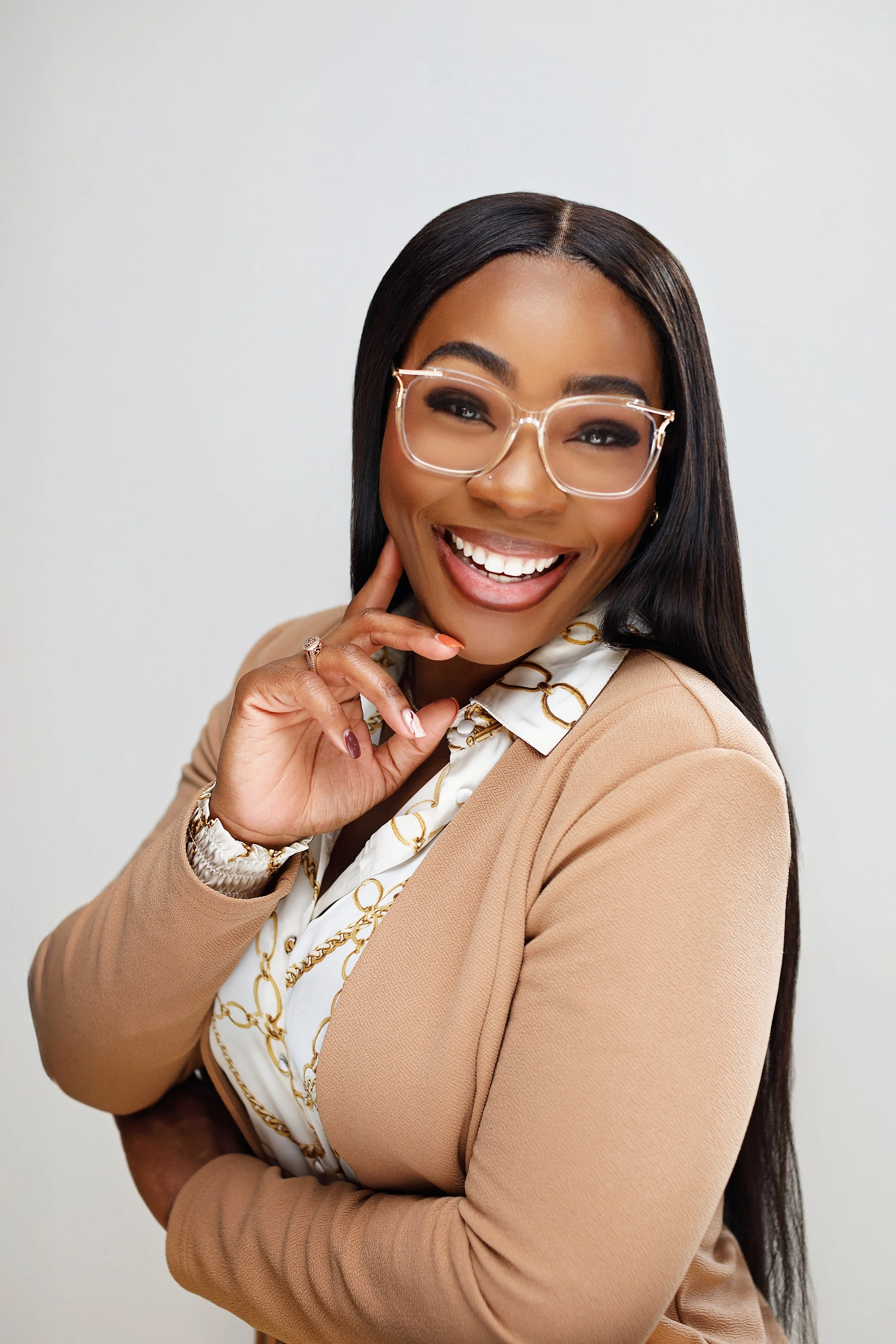 A woman smiling confidently, wearing glasses, a beige blazer, and a white blouse with a gold chain pattern.
