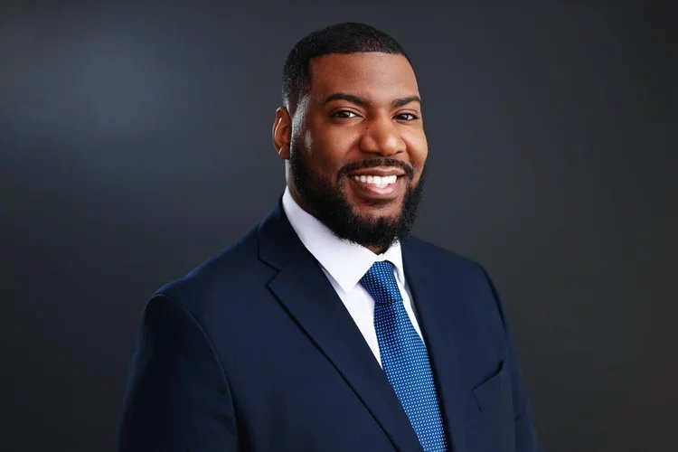 Professional man in a navy blue suit, white shirt, and blue tie smiling against a dark gray background.