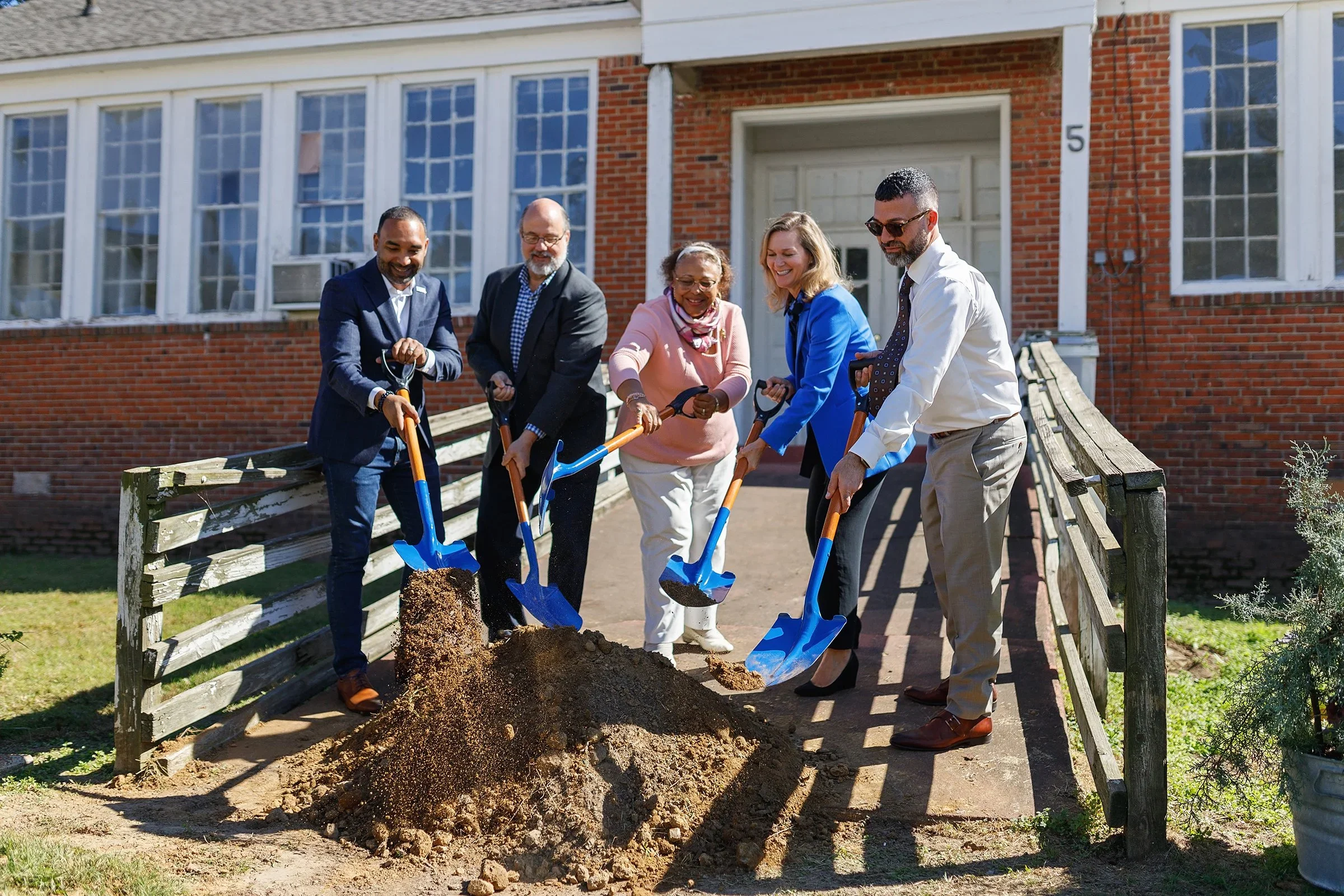 Six people are planting a tree in front of a brick house, shoveling dirt from a hole.