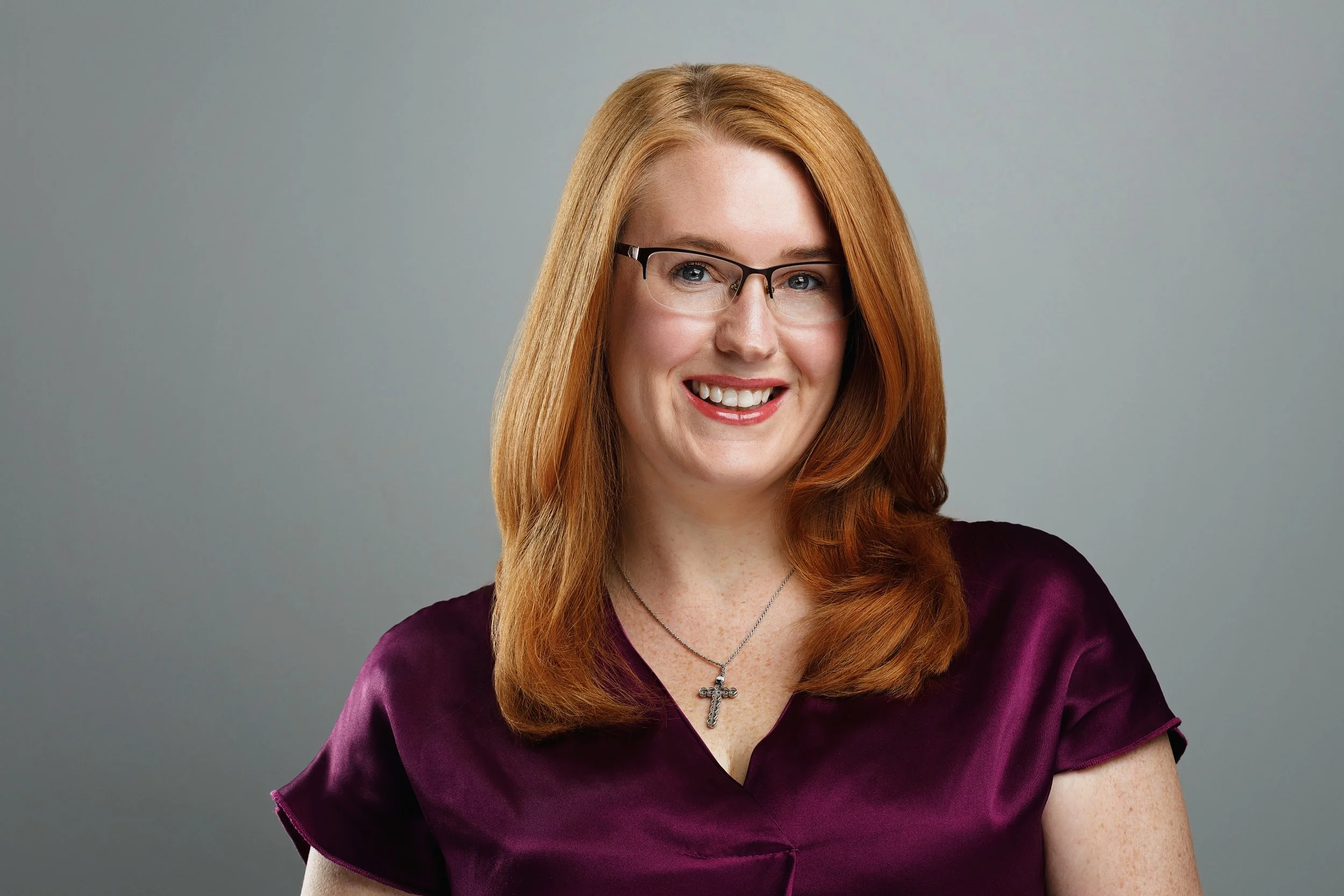 A woman with red hair, glasses, and a purple top, smiling at the camera against a plain gray background.