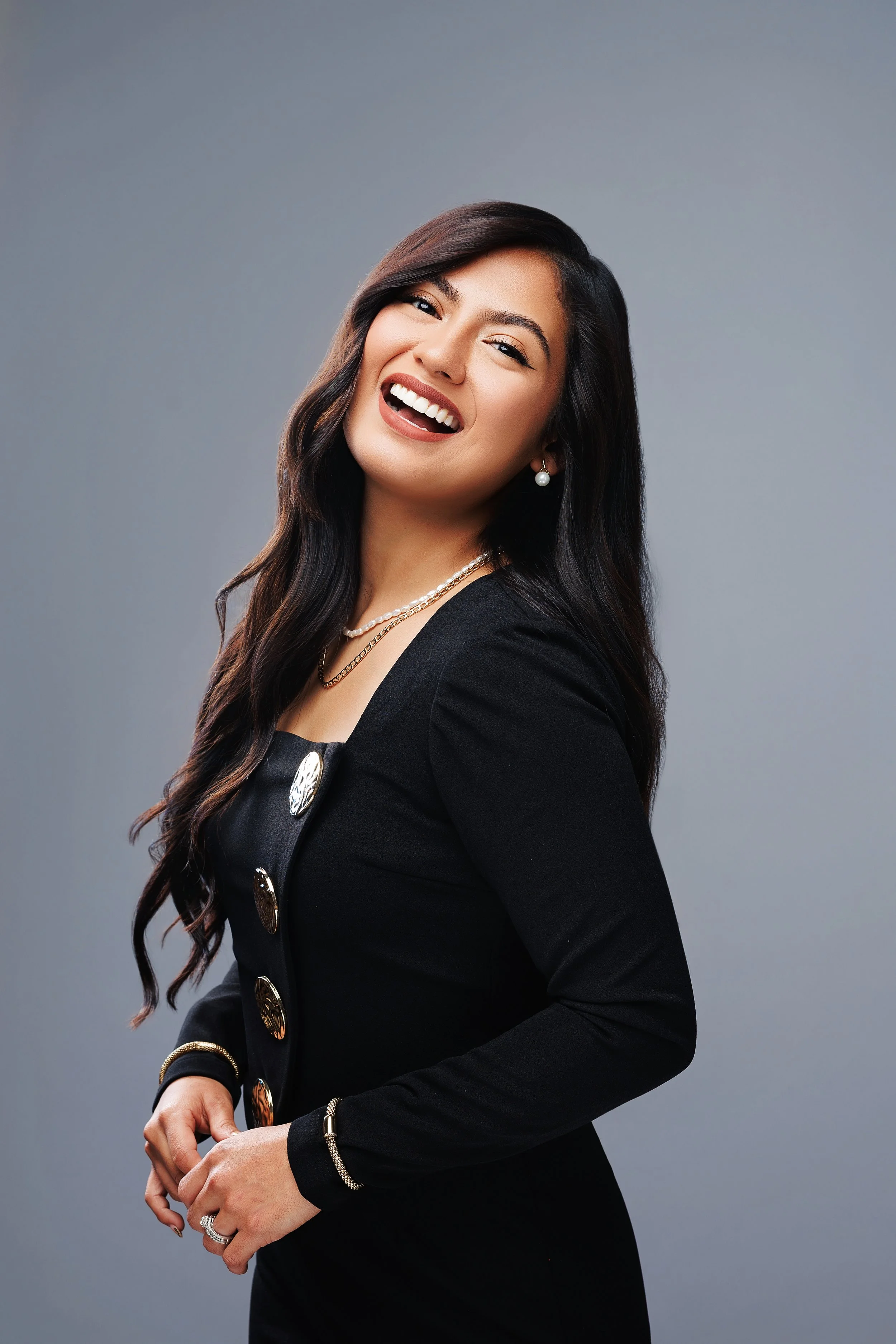 A smiling woman with long dark hair, wearing a black blazer with gold buttons, pearl earrings, and layered necklaces, standing against a gray background.