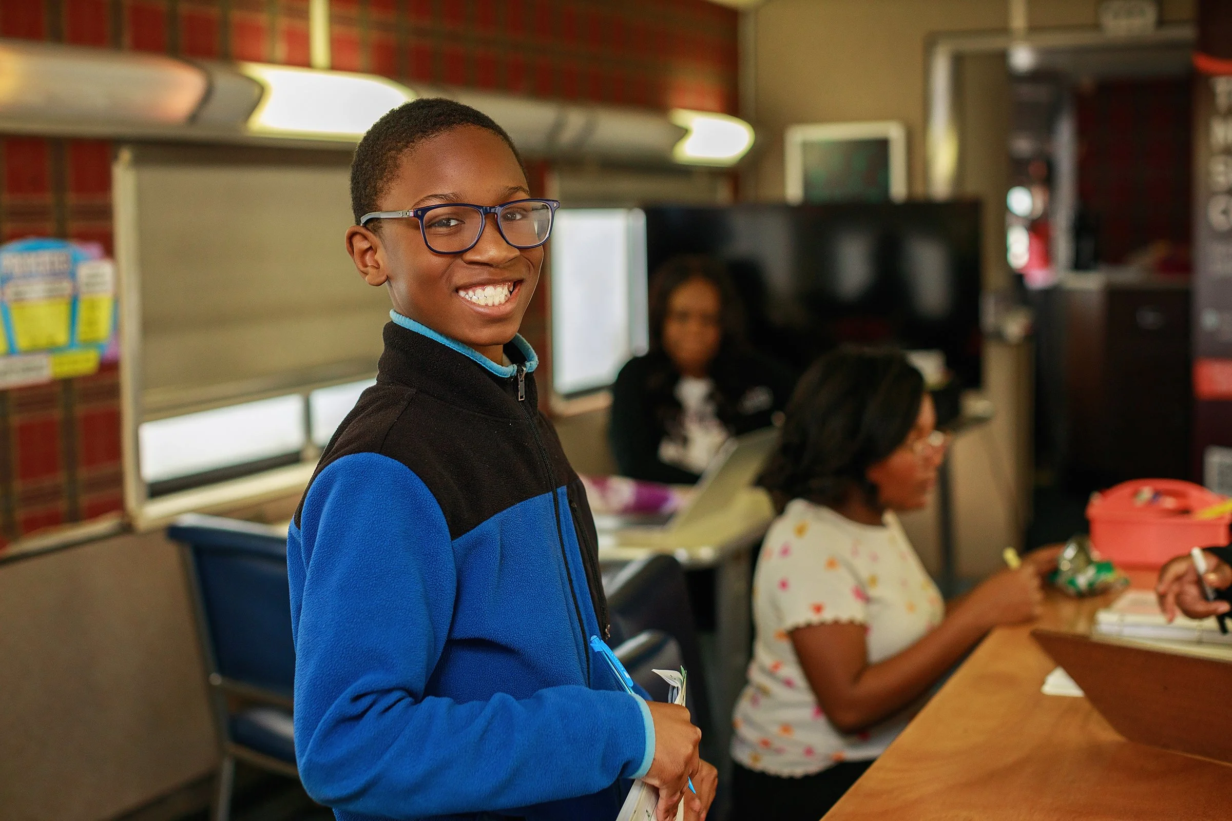 Smiling young boy with glasses and a blue and black jacket in a classroom with two women working at a table in the background.