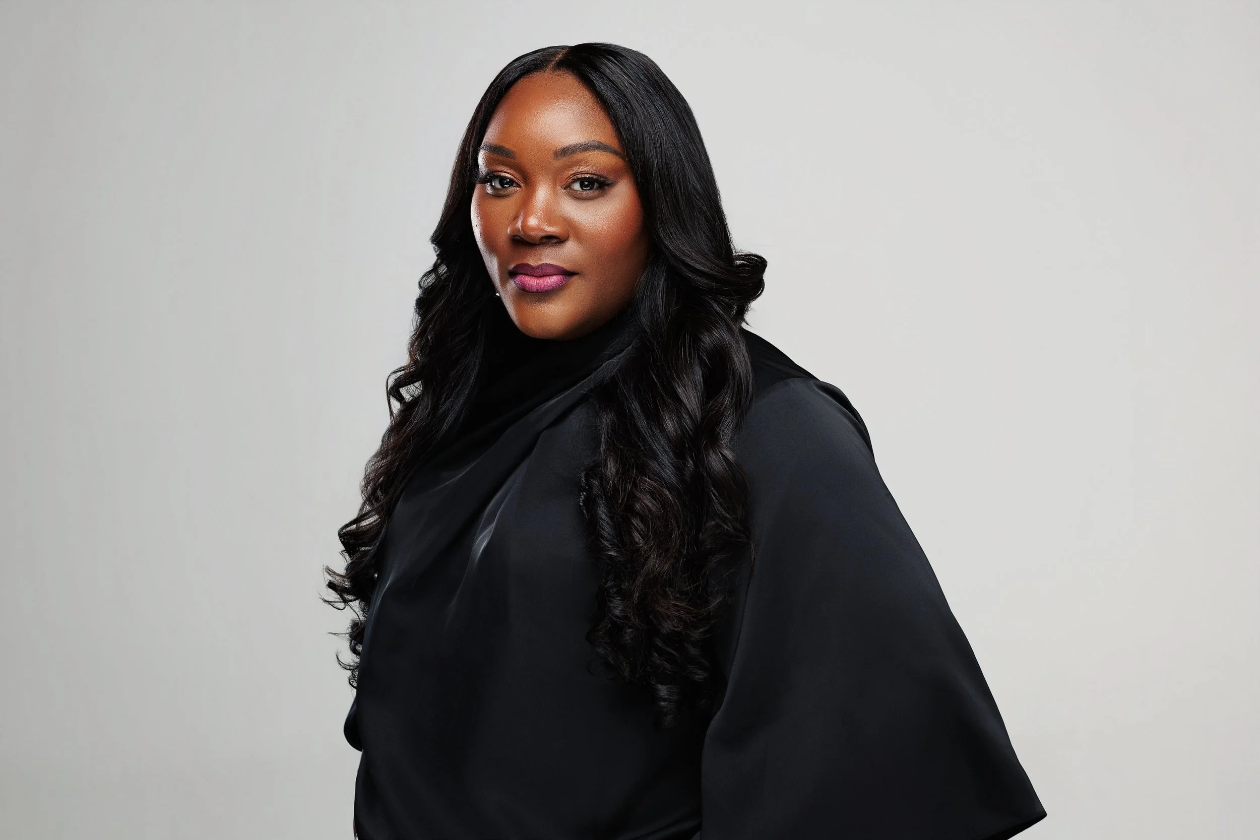 A woman with long black hair wearing a black outfit, looking confidently at the camera against a plain gray background.