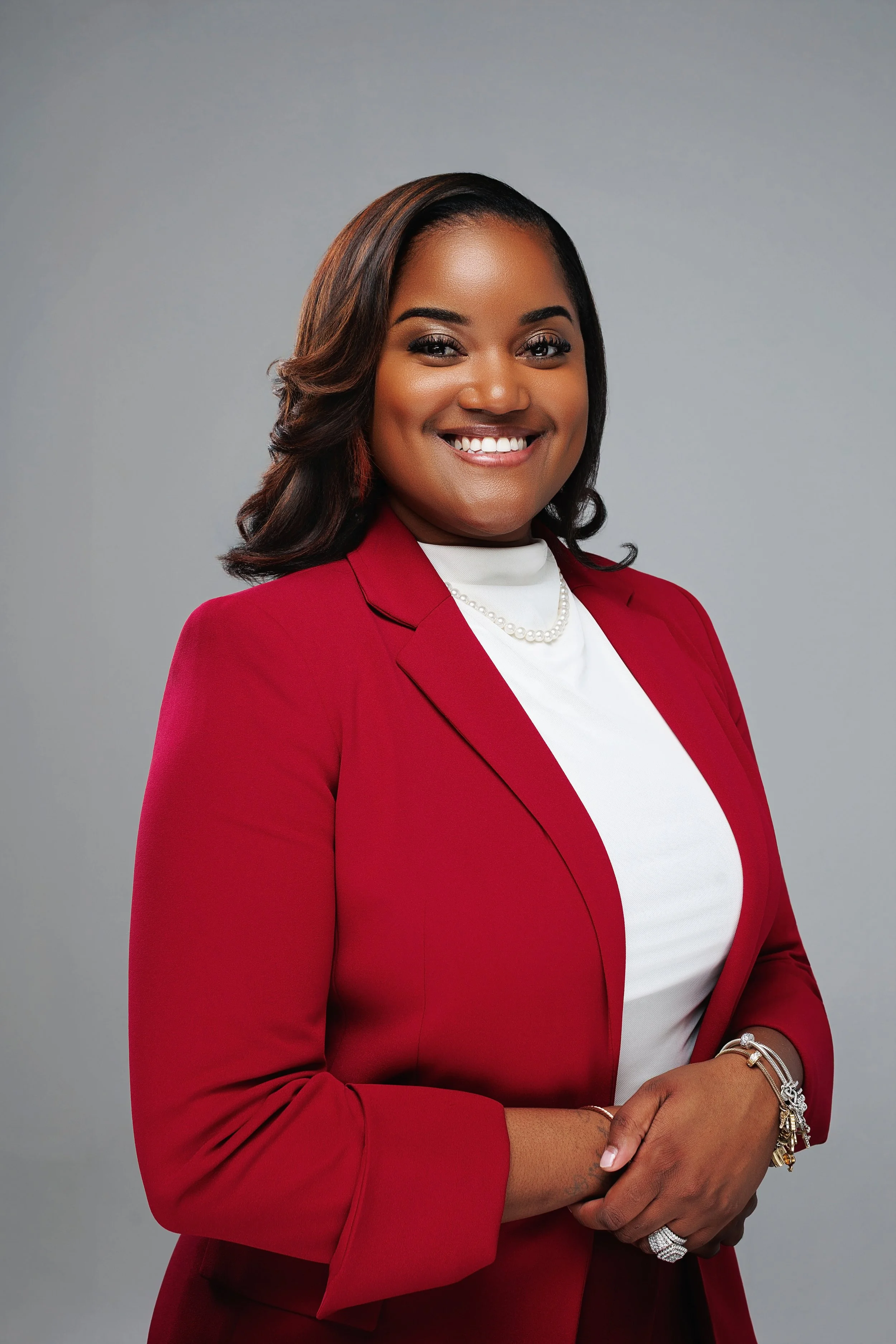A smiling African American woman in a red blazer, white top, and jewelry, posing against a plain gray background.
