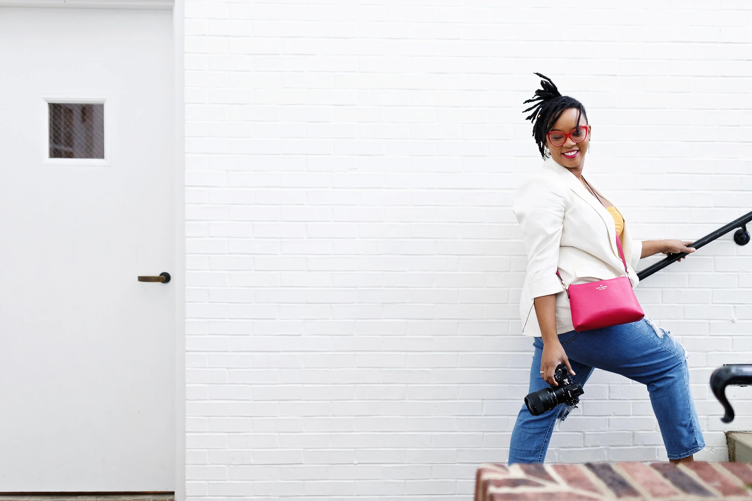 A woman with dreadlocks wearing red glasses, a white blazer, yellow top, and ripped jeans, smiling while holding a camera in one hand and a pink purse in the other, standing on a stairway against a white brick wall.
