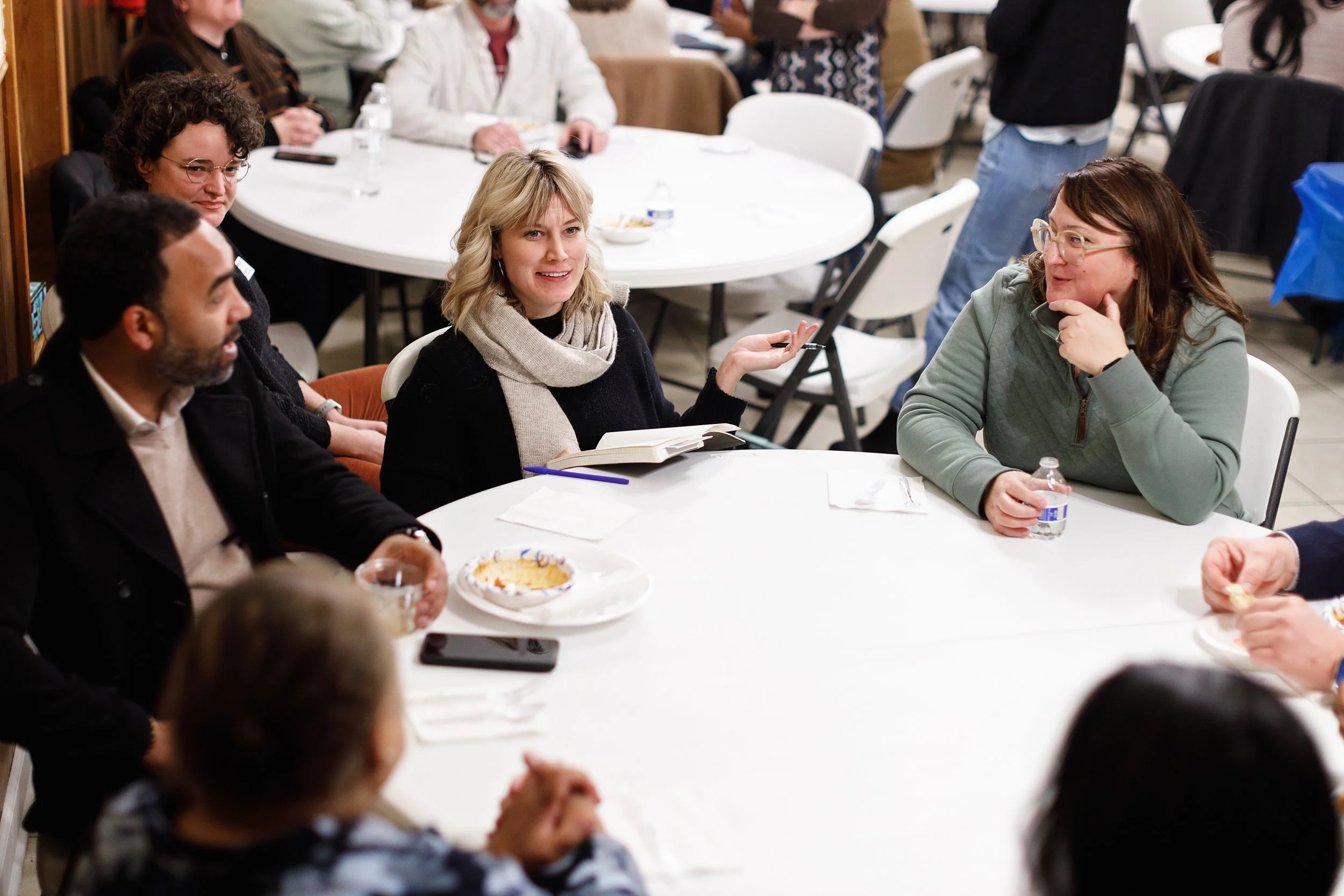 People sitting around a white table, engaged in conversation at a social gathering or meal, with plates, water bottles, and notebooks visible.
