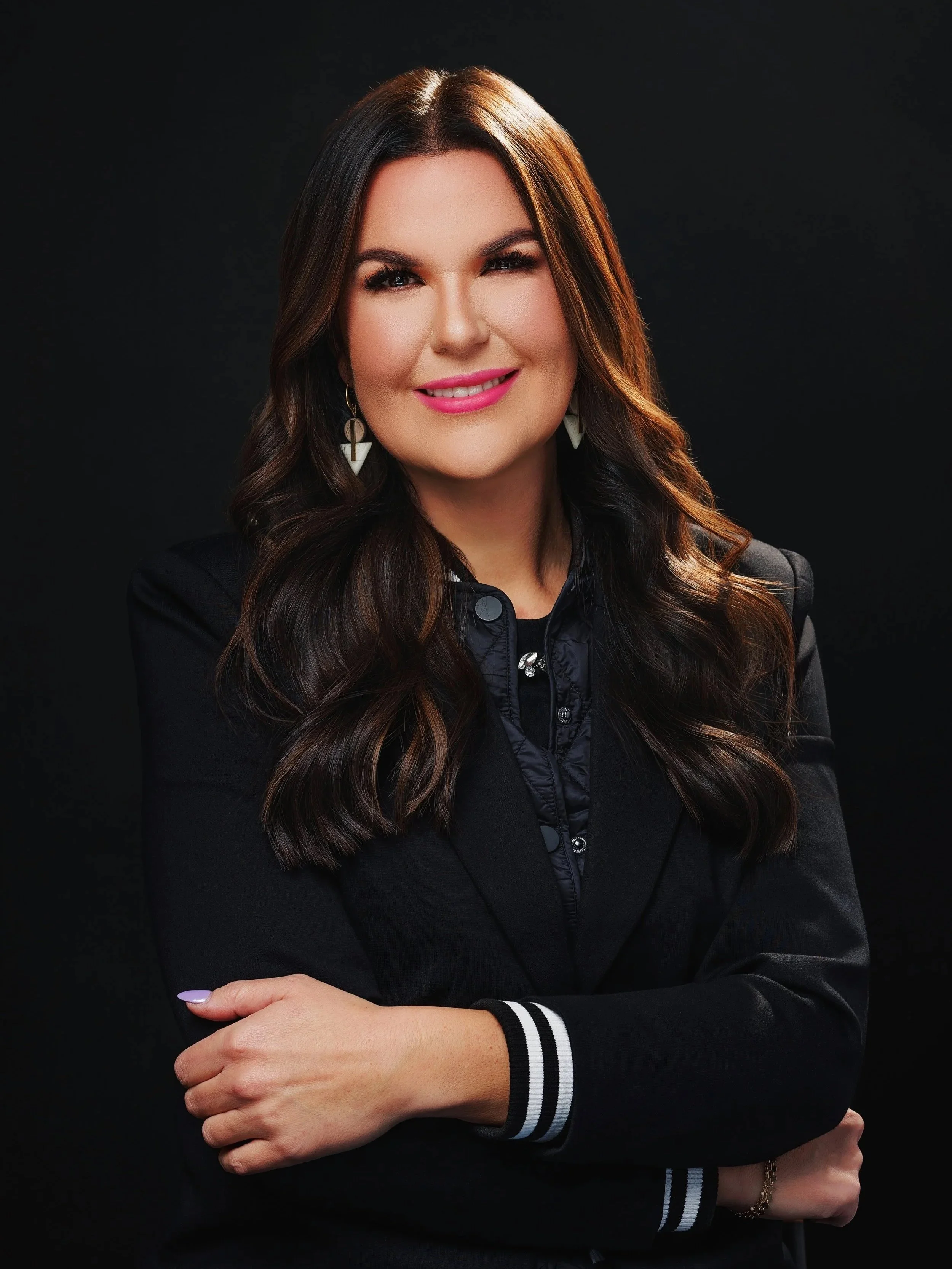 A woman with long brown hair and pink lipstick smiling, wearing a black blazer with striped cuffs, a black top with pearl and rhinestone accents, and earrings, against a solid dark background.