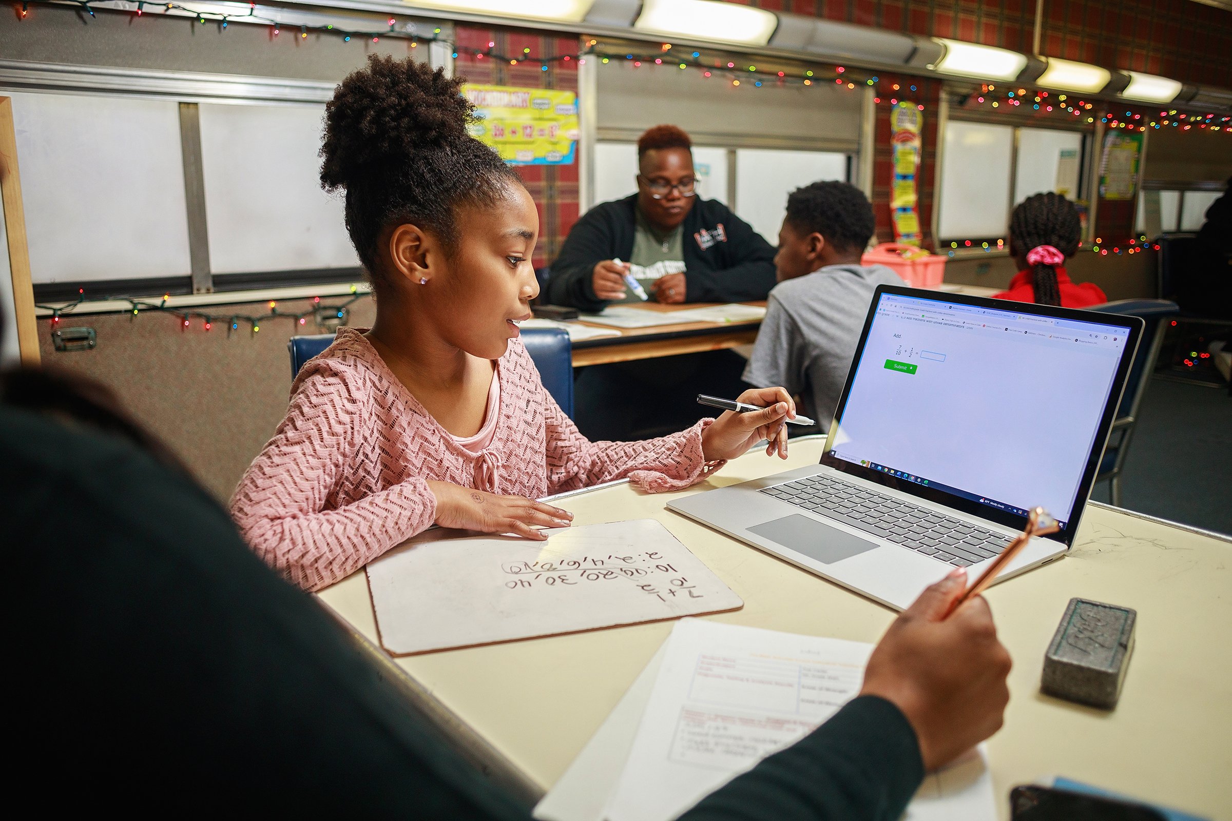 A young girl in a pink sweater studies math problems on a whiteboard while working on an online assignment on her laptop. An adult's hand holding a pencil is nearby, and in the background, other students and a woman are seated at table in a decorated