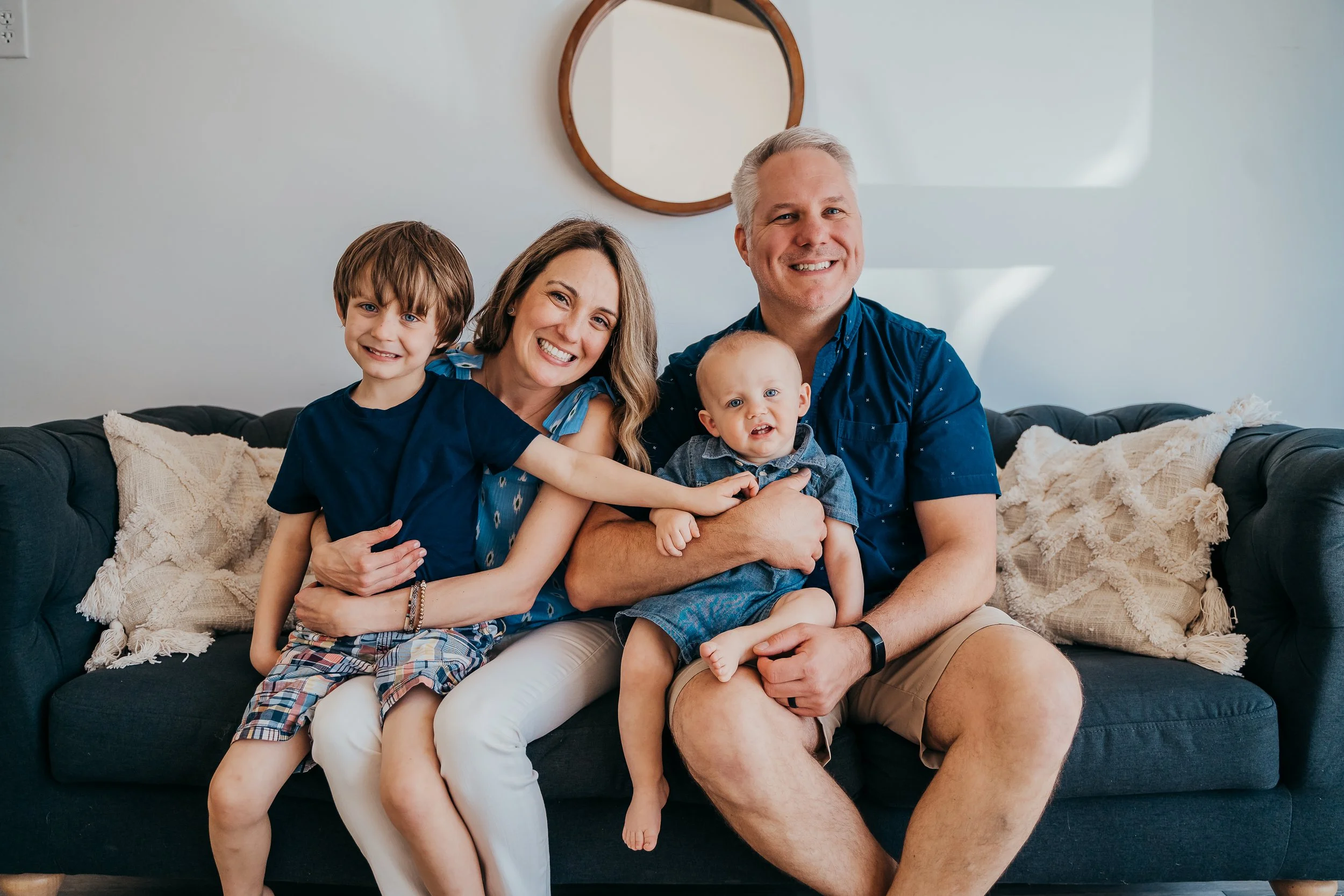 A happy family sitting on a dark sofa in a living room, with a circular mirror on the wall behind them. The family includes two adults and two young children, all smiling and dressed in casual blue and denim clothing.