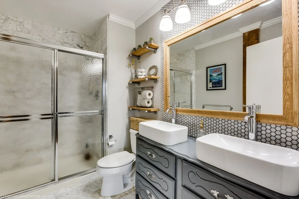 A modern bathroom with a glass shower, a white toilet, a double sink vanity with black drawers, and a large mirror with a wooden frame. Open wooden shelves hold towels and decor, and there is a framed poster on the wall.