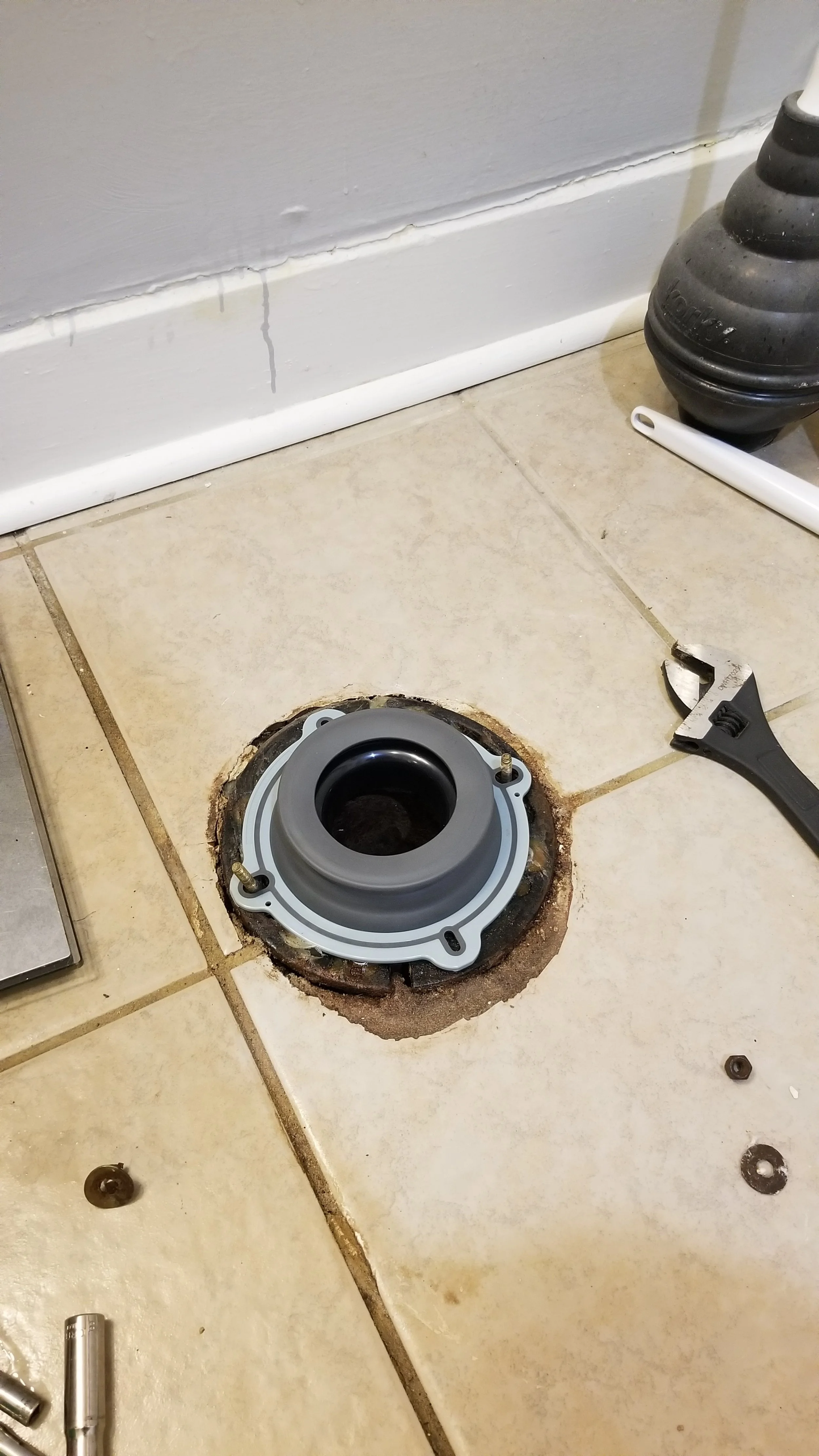 A bathroom floor with a removed toilet flange, revealing the pipe opening and surrounding area. Tools such as an adjustable wrench and screws are nearby, and tiles are partially removed around the flange.