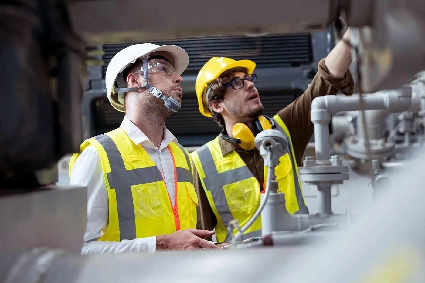 Two male industrial workers wearing yellow safety vests and helmets, working on machinery in an industrial setting.