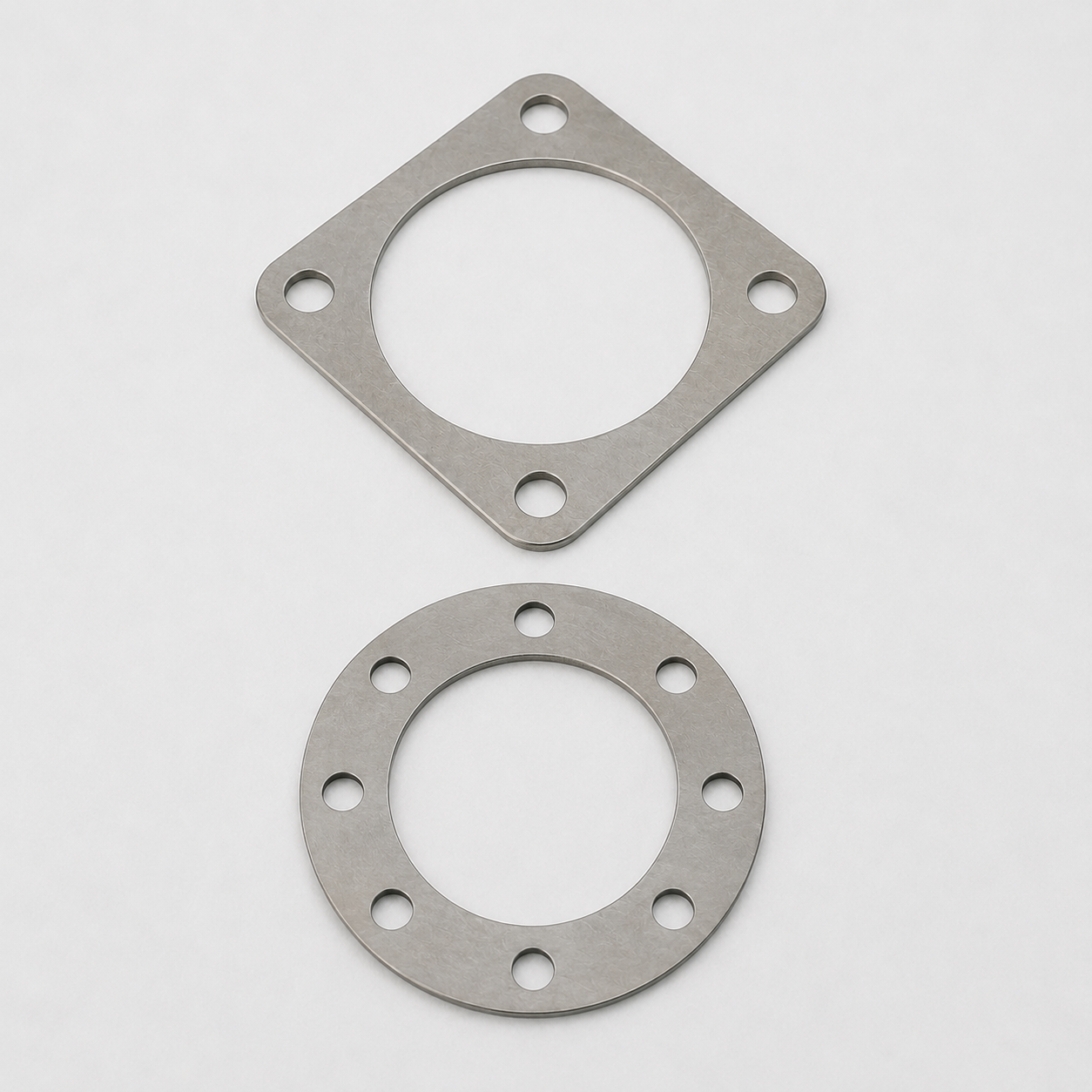 Two metallic gaskets, one square and one round, with holes around their edges, placed on a plain white background.