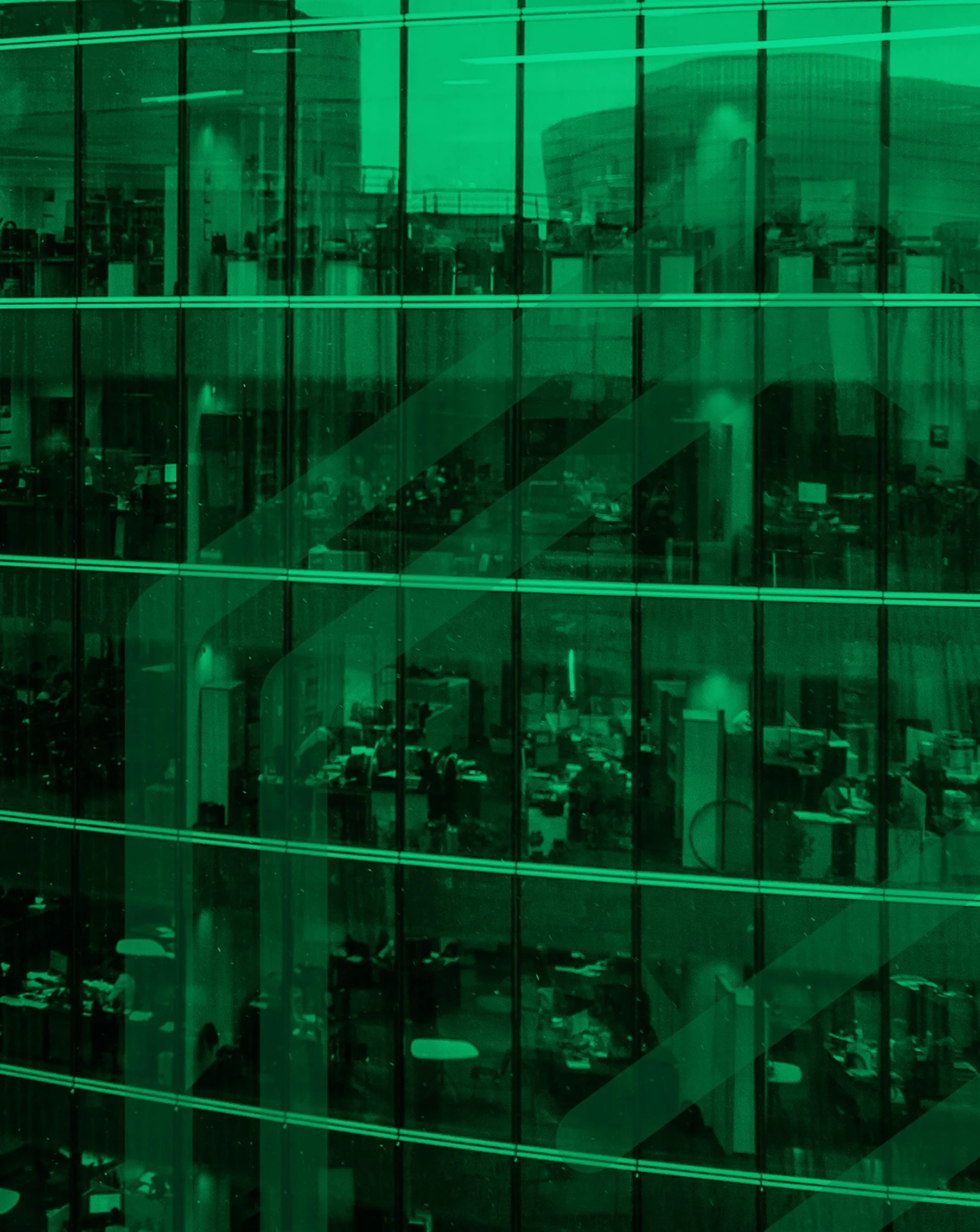 View of the interior of a modern office building through green-tinted glass windows, showing desks, chairs, and office equipment inside.