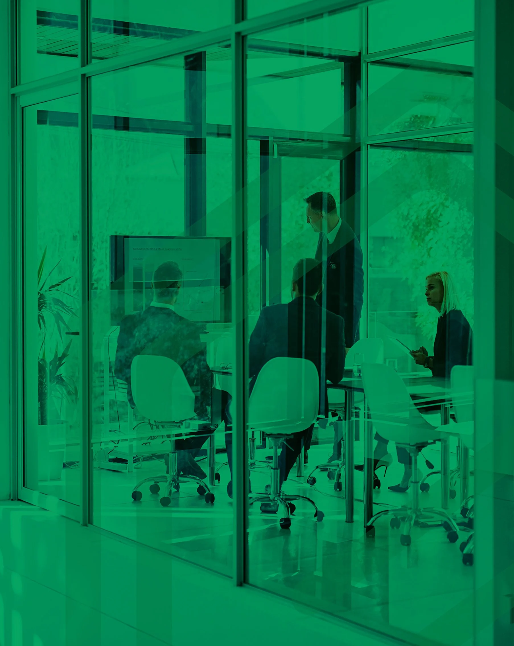 An office conference room with glass walls and chairs around a table, with a man standing and a woman sitting, all engaged in a discussion.