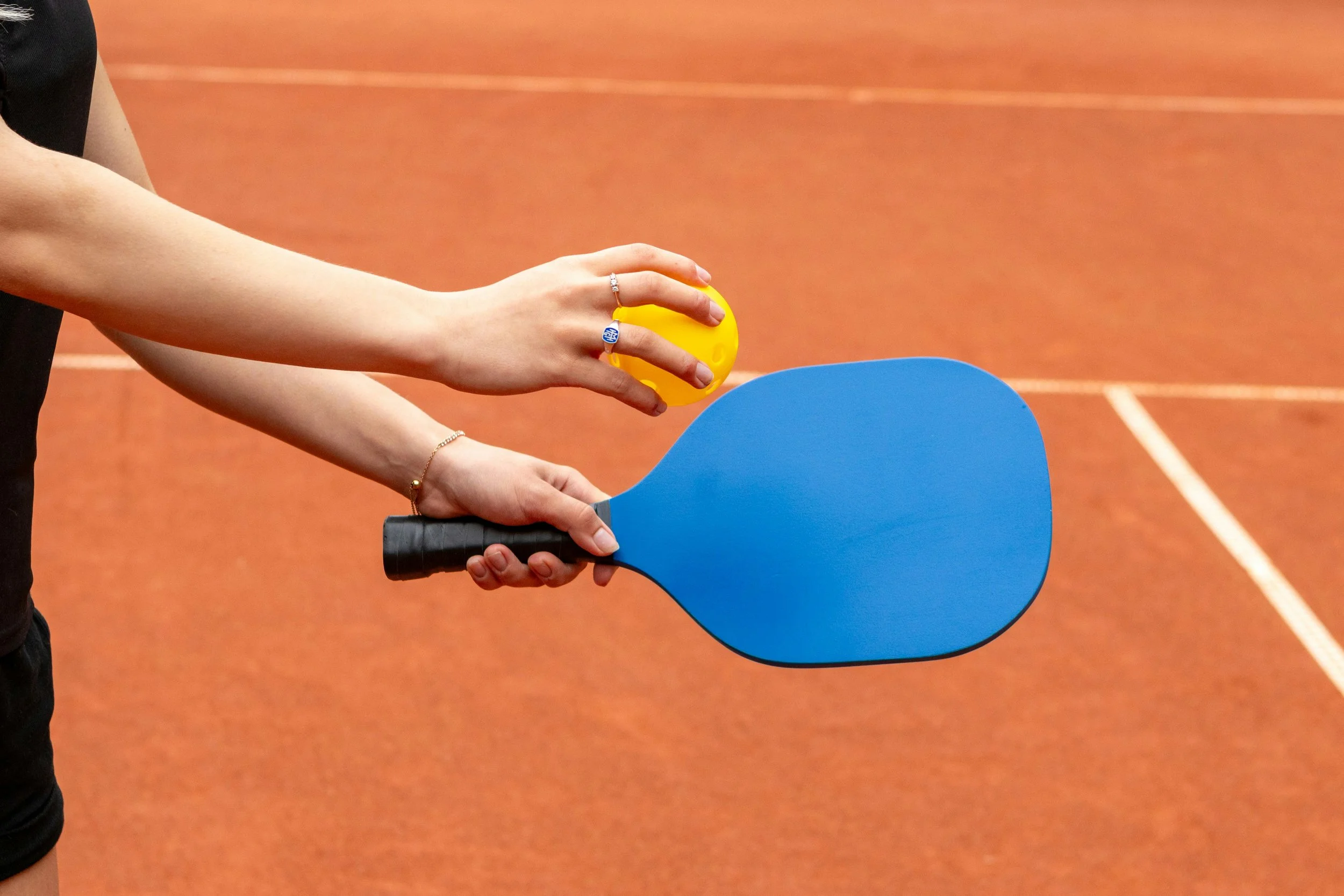 Person holding a blue ping pong paddle and a yellow ping pong ball on an orange indoor court.