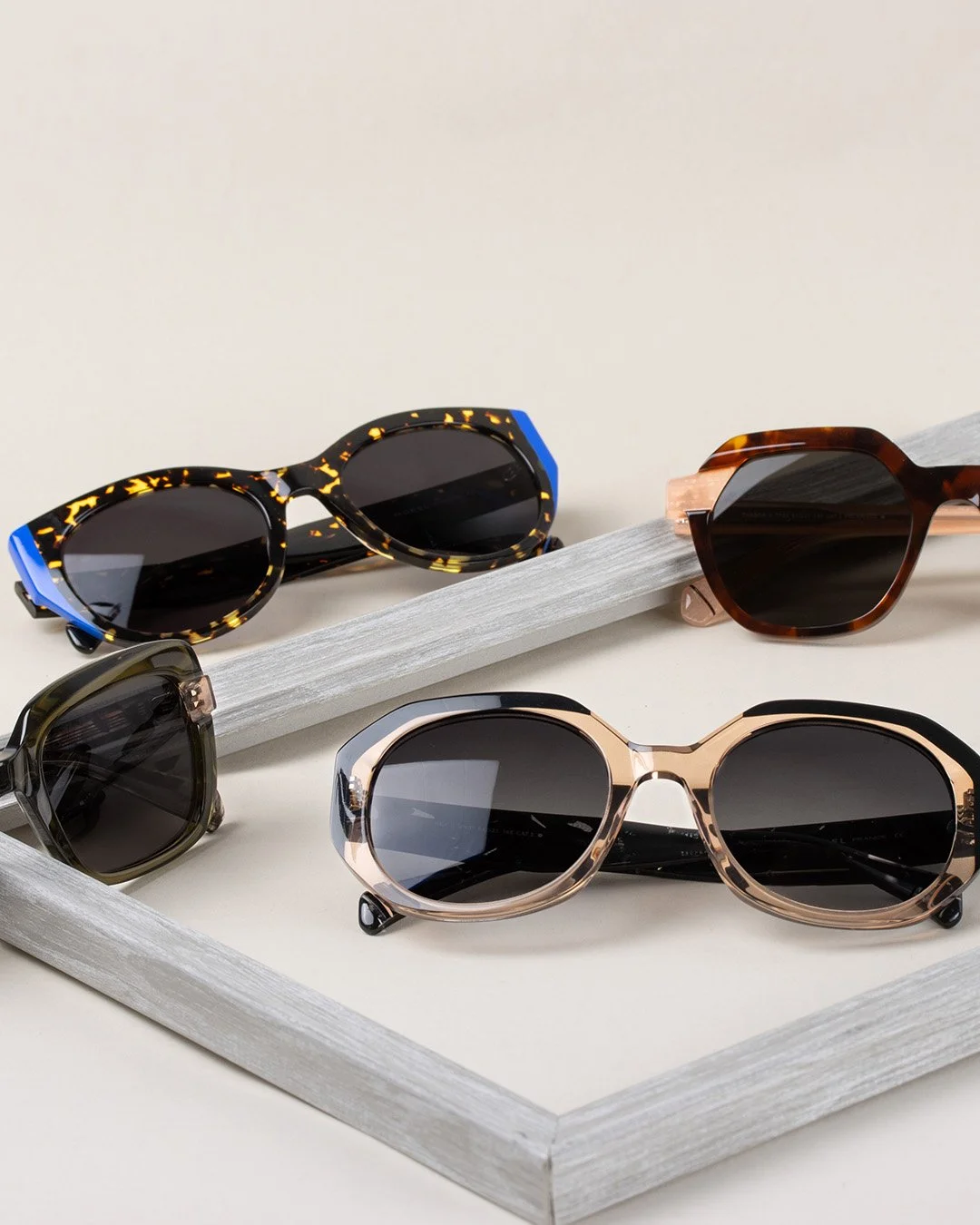 Four pairs of stylish sunglasses with different frame designs and colors displayed on a white wooden frame against a plain background.
