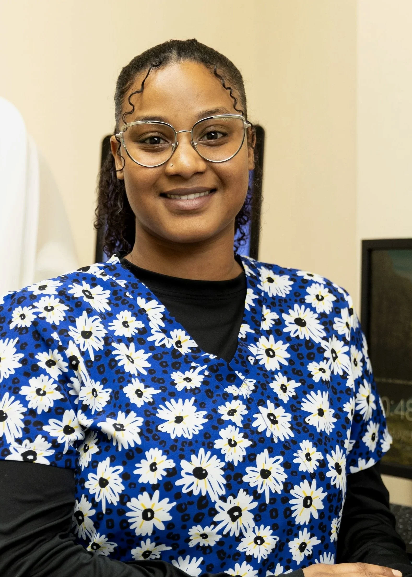 A woman standing indoors, wearing glasses and a blue floral scrub top, smiling at the camera.